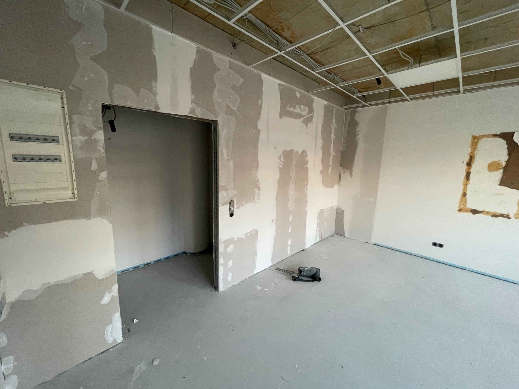Pokój w trakcie szpachlowania ścian, widoczne ślady po łatach gipsowych, sufit z instalacją, zabezpieczona podłoga i narzędzie do szpachlowania.