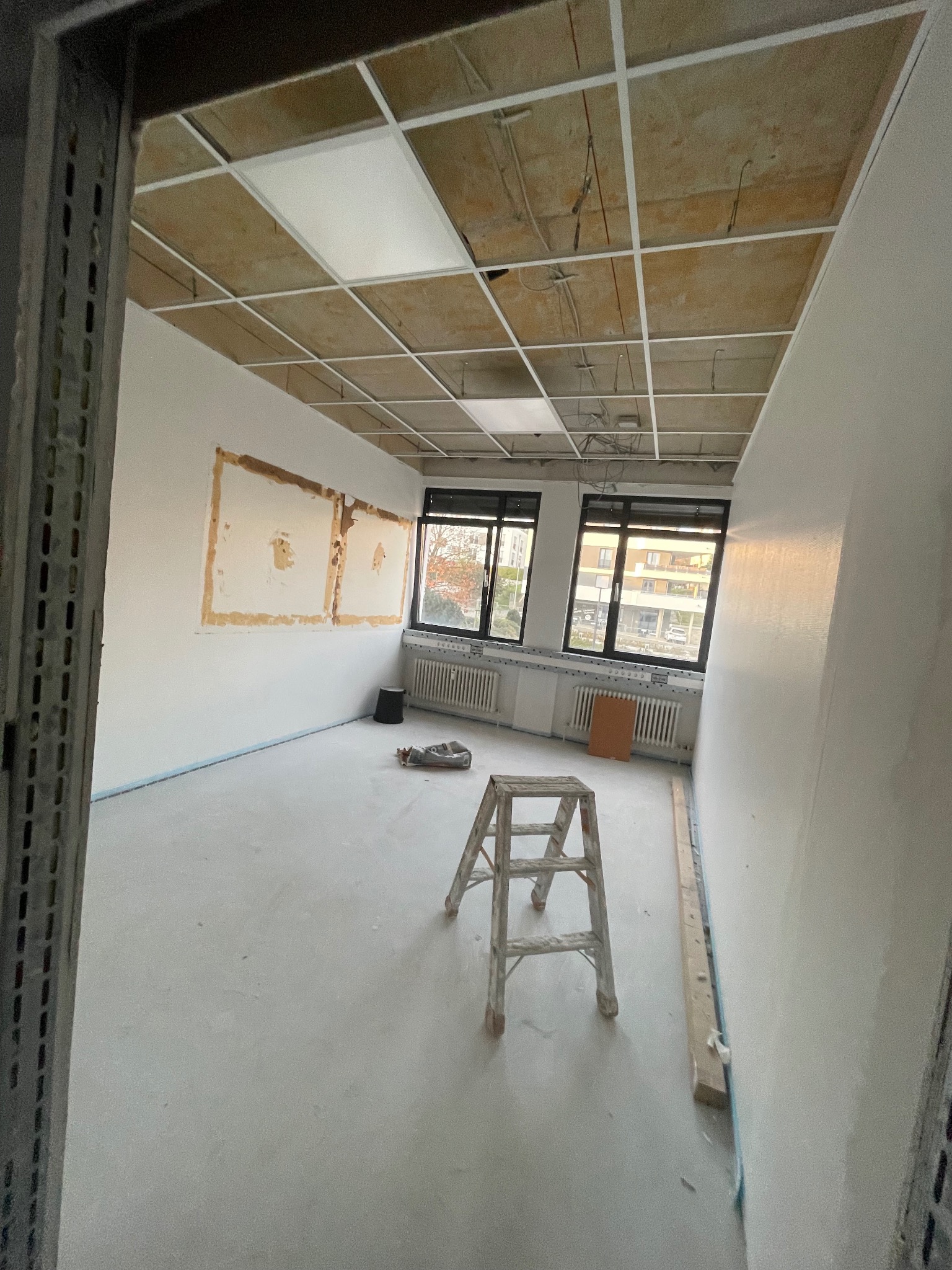 Wnętrze w trakcie prac: sufit podwieszany w trakcie montażu, ściany przygotowane do malowania, drabina w centrum. Widok zza metalowej framugi.