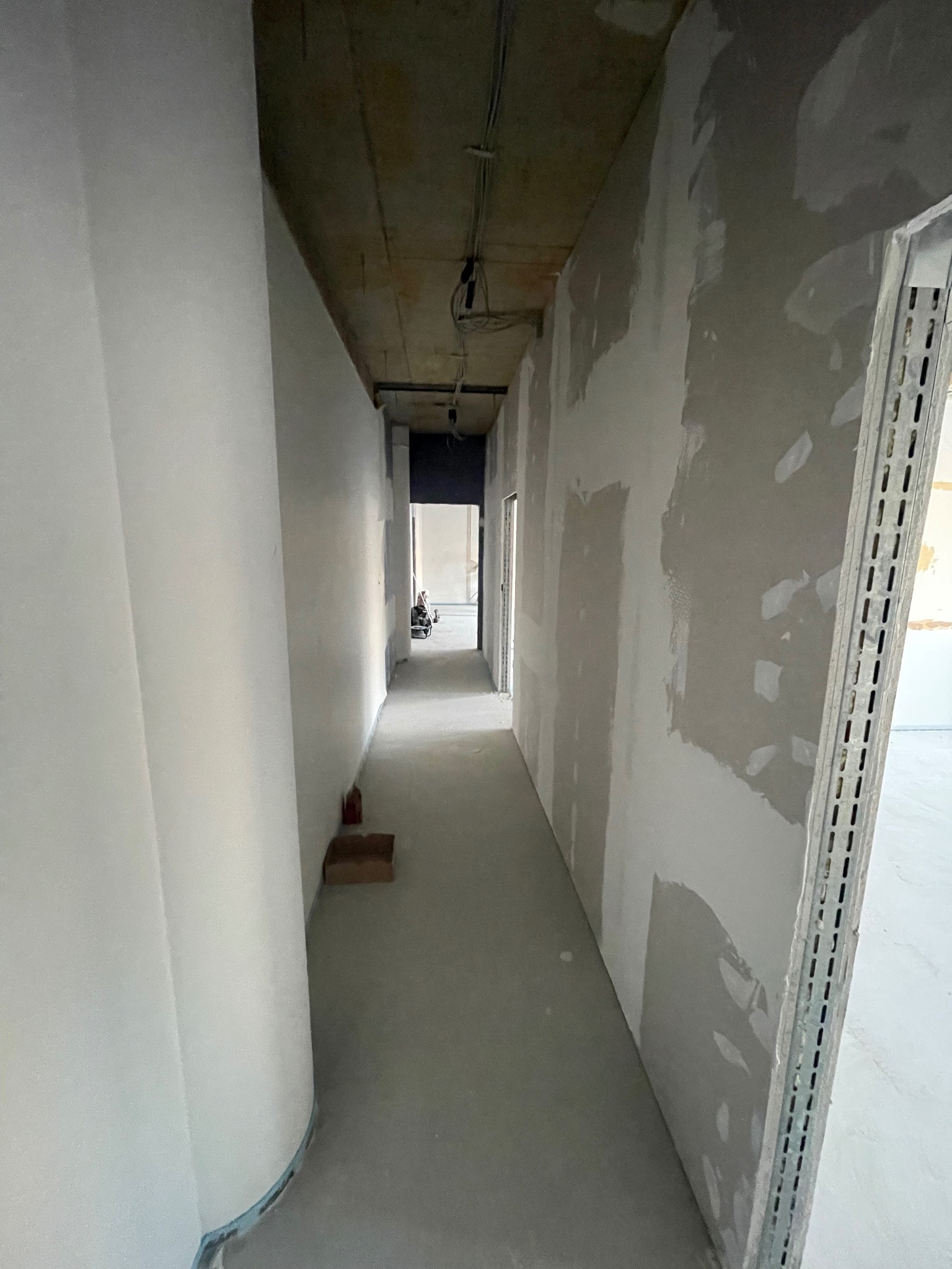 Korytarz w trakcie prac wykończeniowych: ściany częściowo wyszpachlowane, surowy betonowy strop, widoczne profile montażowe. W tle majster przy pracy.