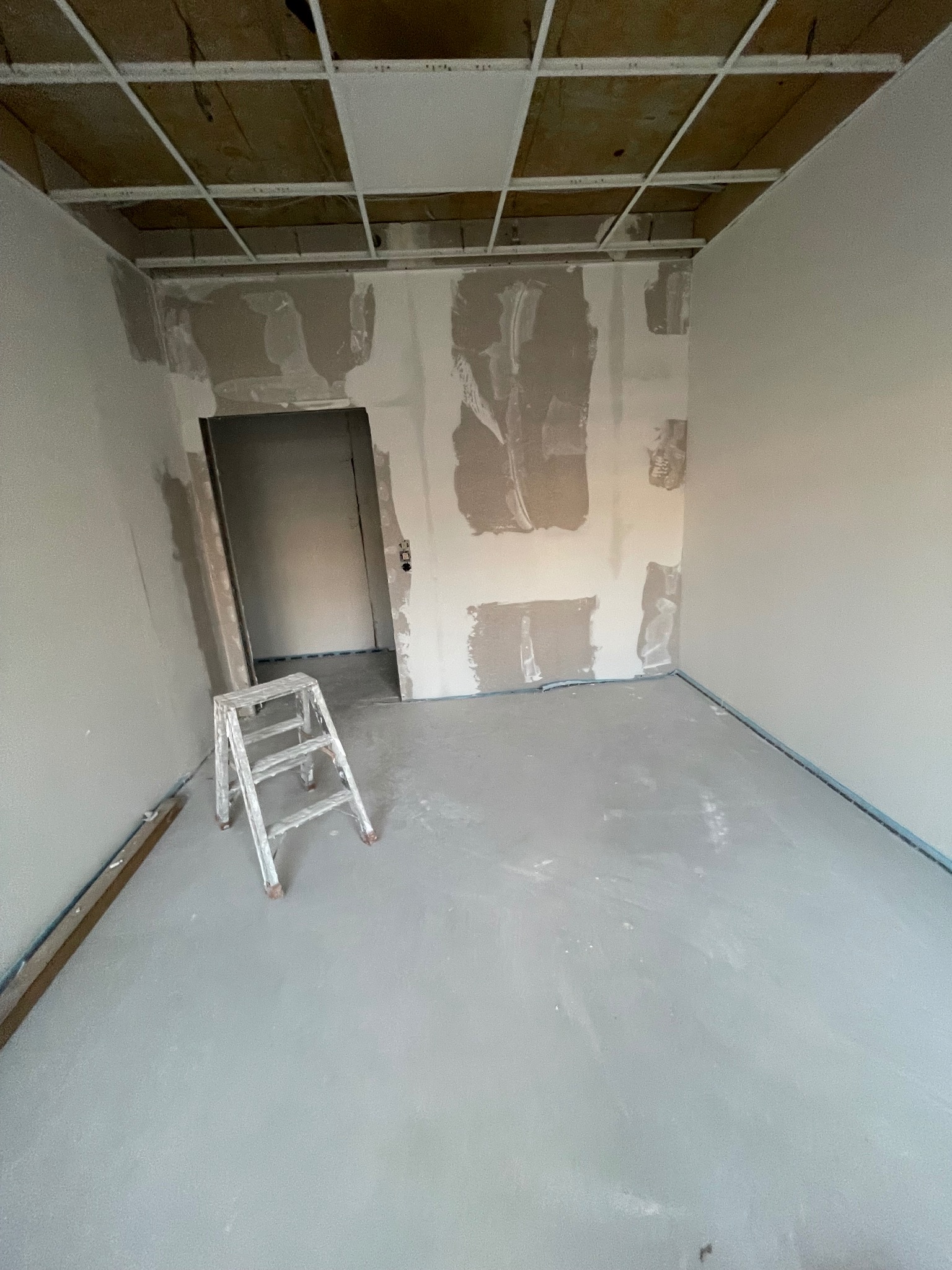 Pokój w trakcie prac wykończeniowych: szpachlowane ściany, sufit z widoczną konstrukcją, drabina malarska i listwa przypodłogowa. Widoczne ślady po pracach na podłodze.