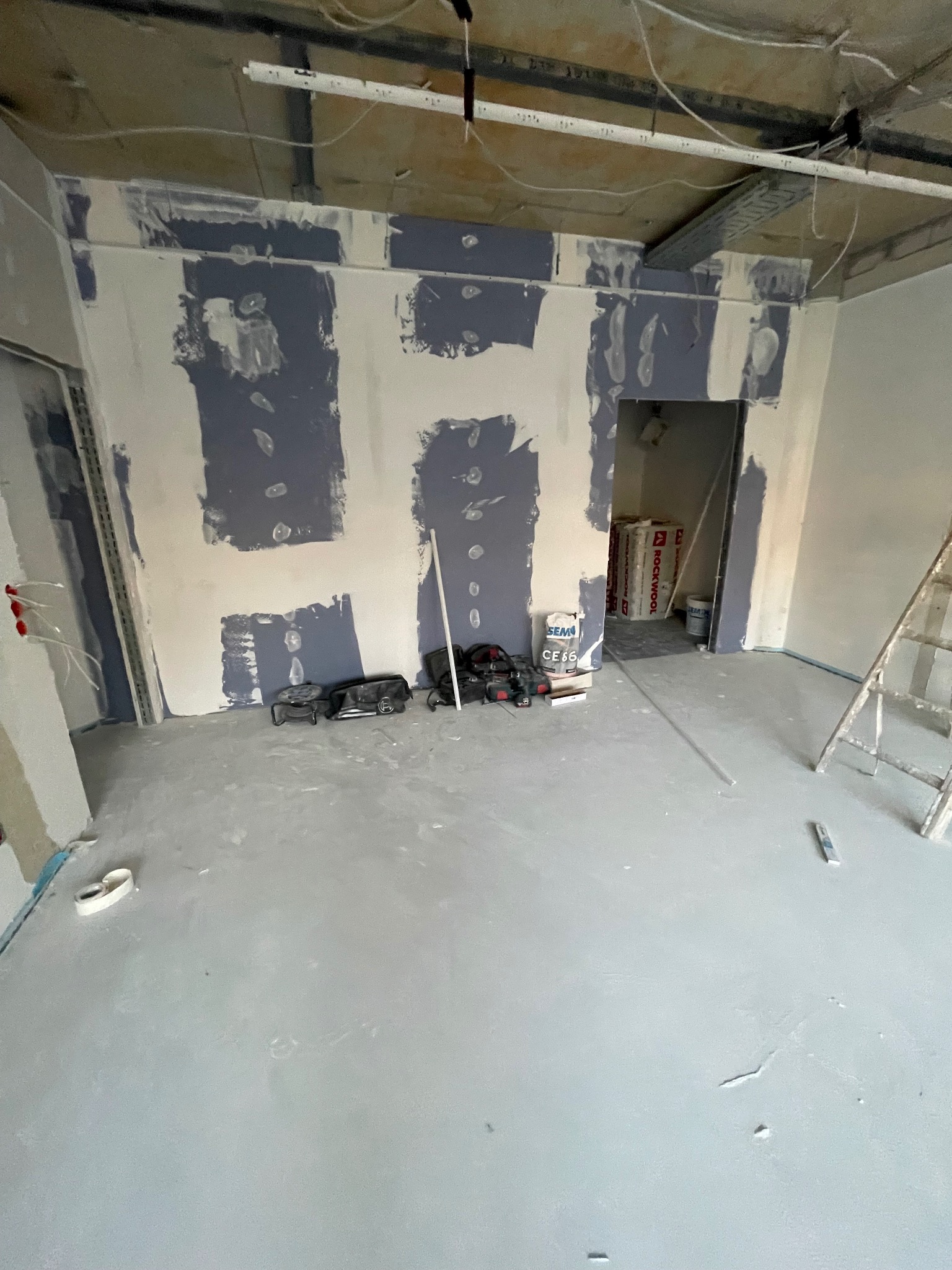 Wnętrze w trakcie prac: ściany z częściowo nałożoną gładzią, narzędzia na podłodze, widoczne instalacje pod sufitem, drabina oparta o ścianę.