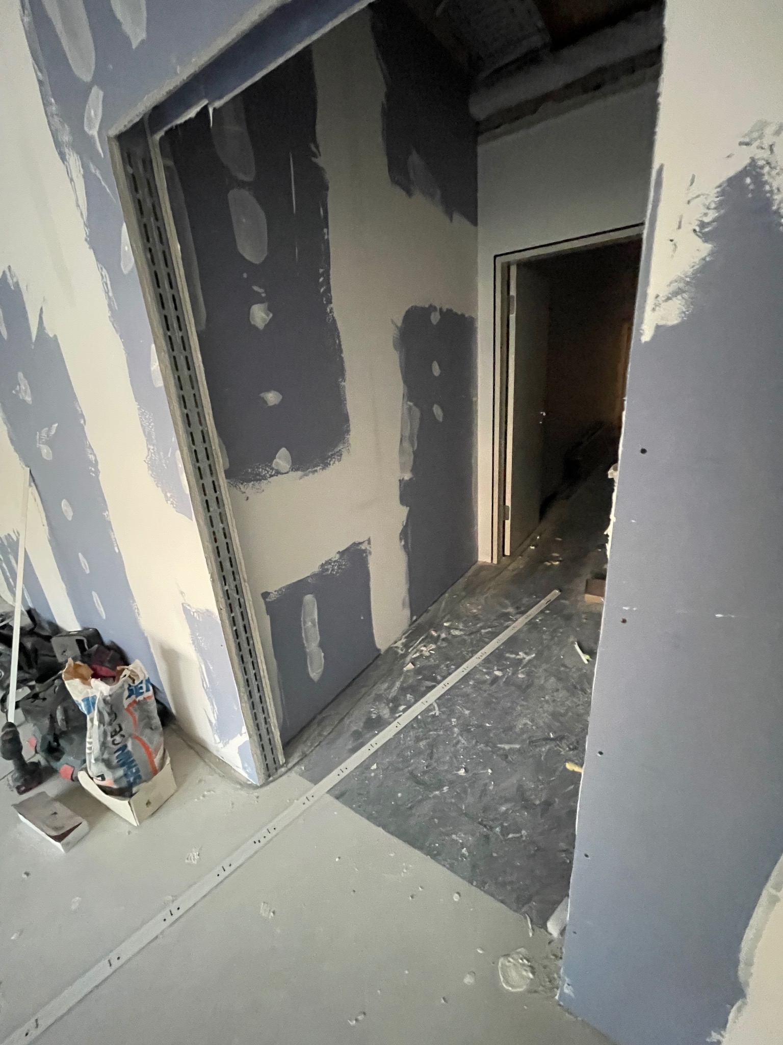 Wnętrze w trakcie prac: ściany z płyt karton-gips, widoczne profile metalowe, szpachlowane fragmenty. Przy podłodze narzędzia i materiały budowlane.