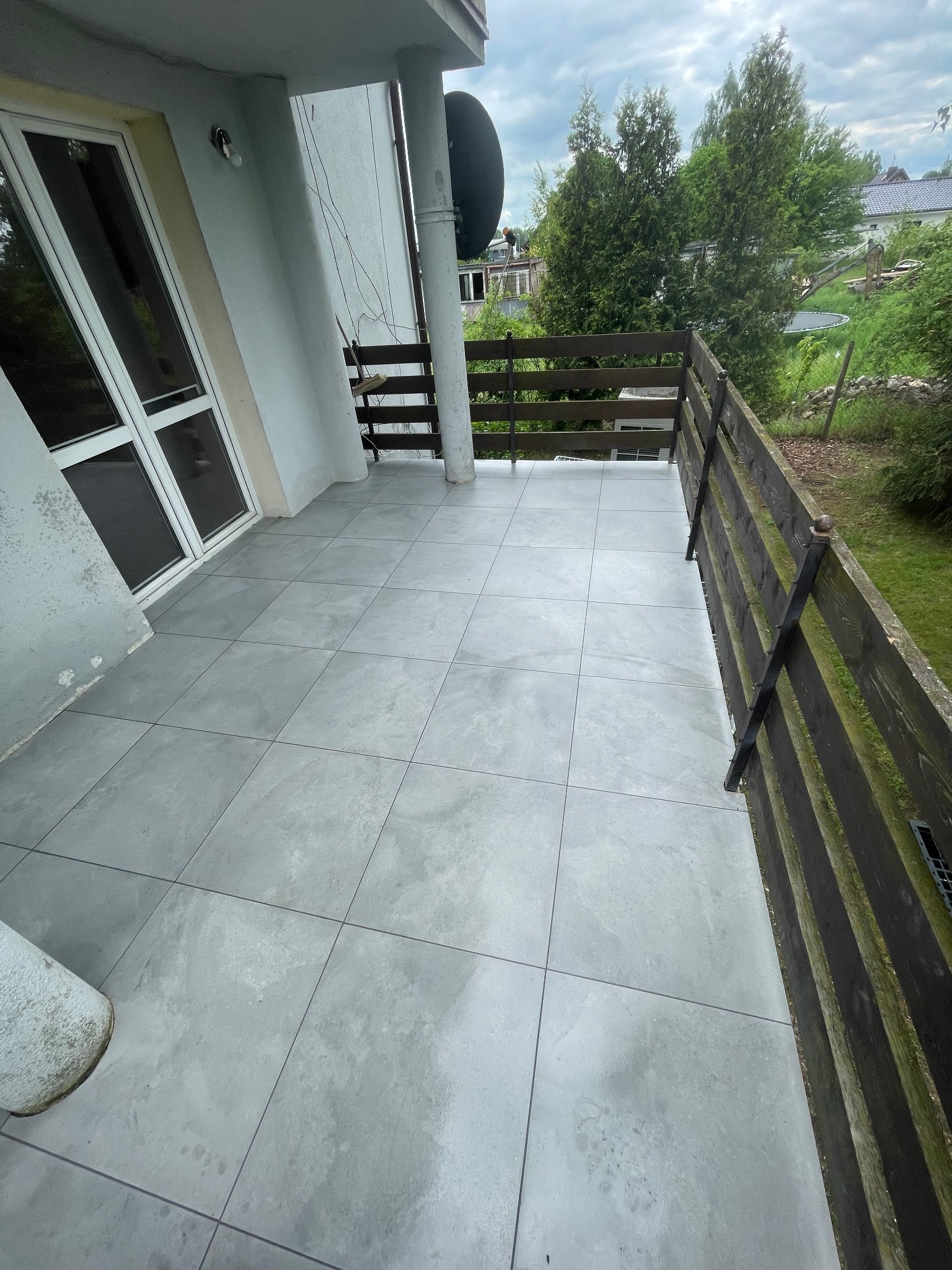 Szare płytki na balkonie z drewnianą balustradą. Widok na zieleń. Nowoczesne wykończenie balkonu z widocznymi fugami i detalami architektonicznymi budynku.