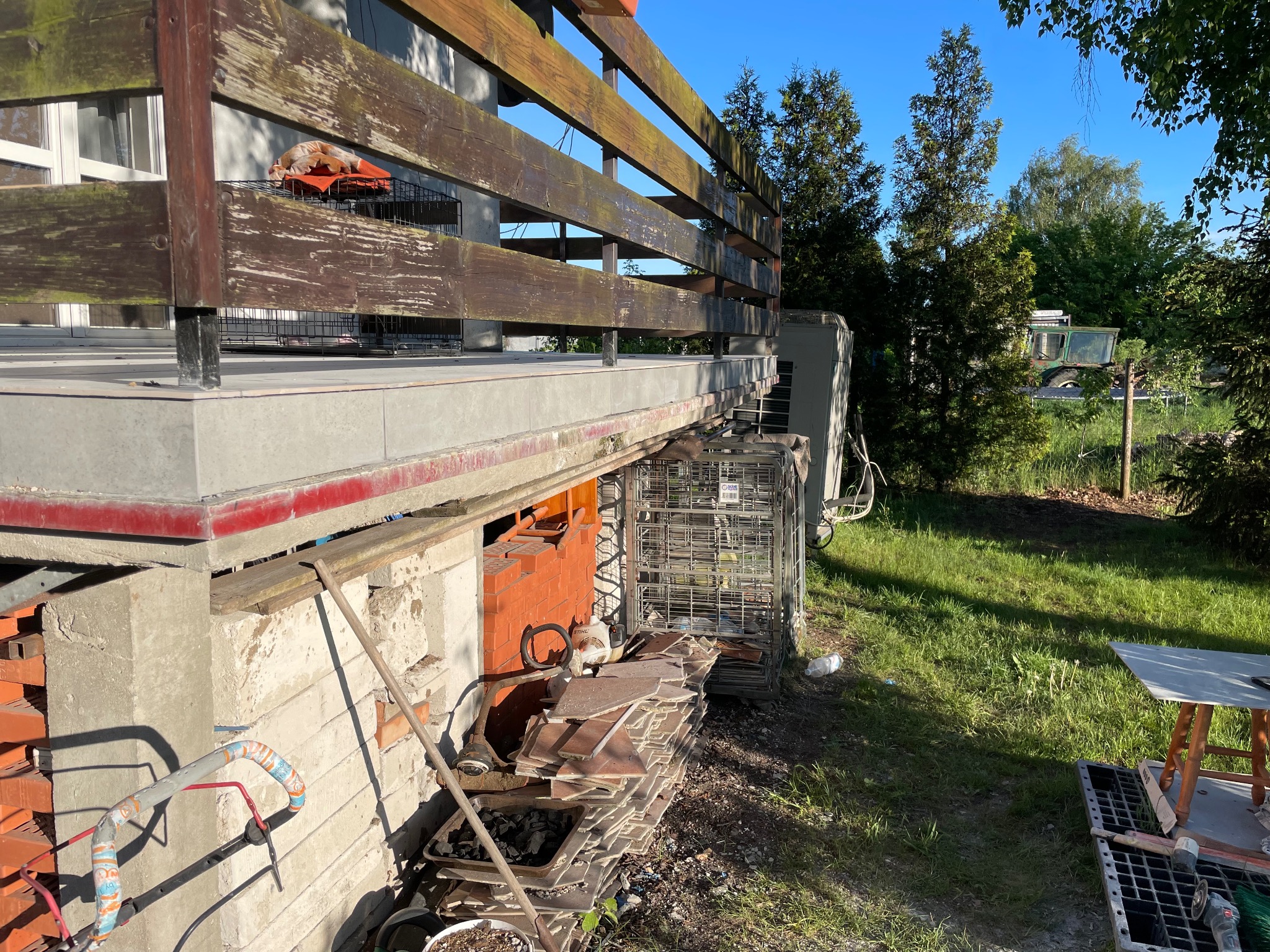 Balkon w trakcie remontu: widoczne cegły, płytki, narzędzia i materiały budowlane. Prace wykończeniowe w toku, zieleń w tle.