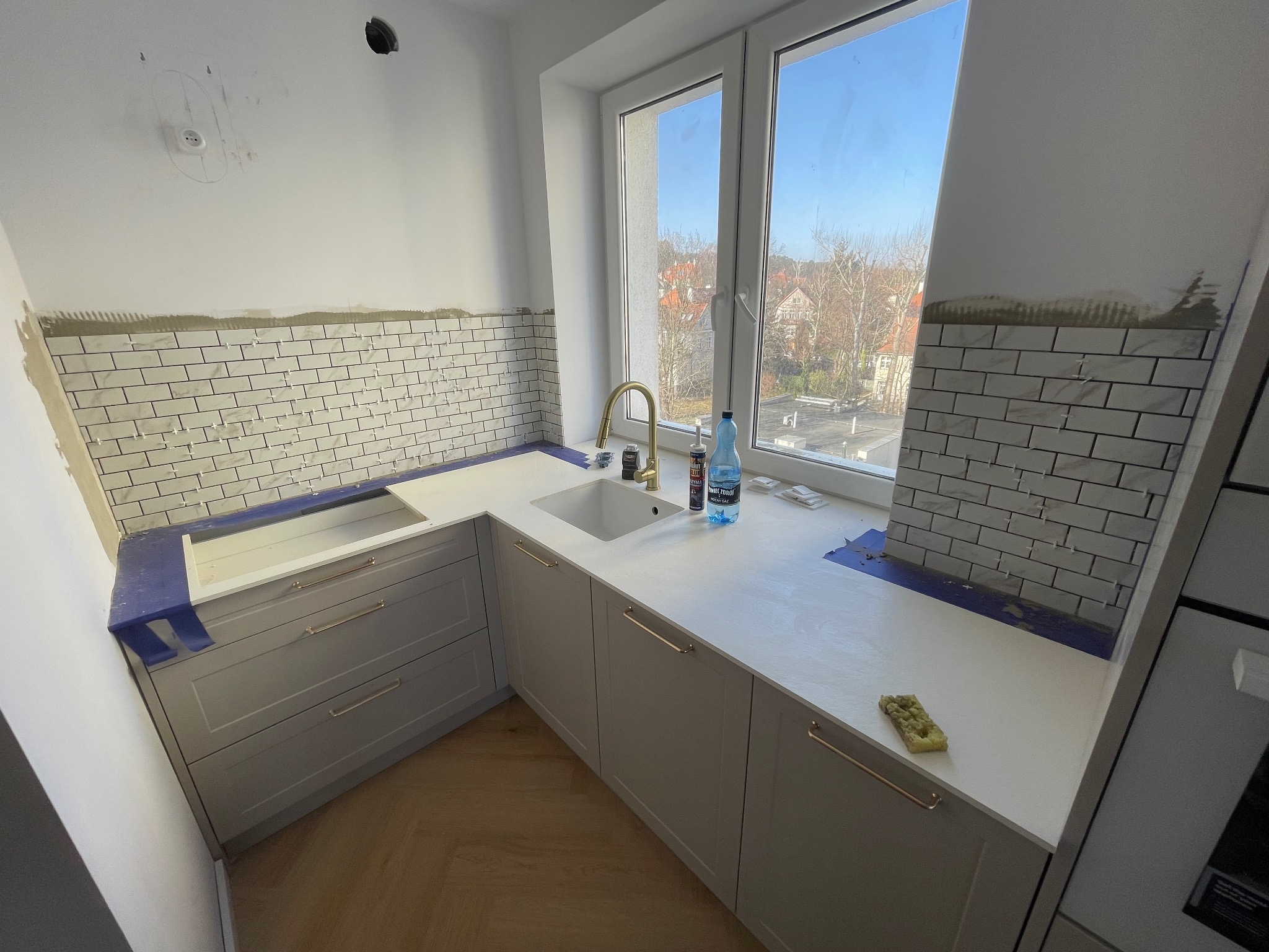 Kuchnia w trakcie wykańczania: białe płytki ułożone do połowy ściany, szare szafki z złotymi uchwytami, biały blat, okno z widokiem na miasto. Widoczne narzędzia i materiały.