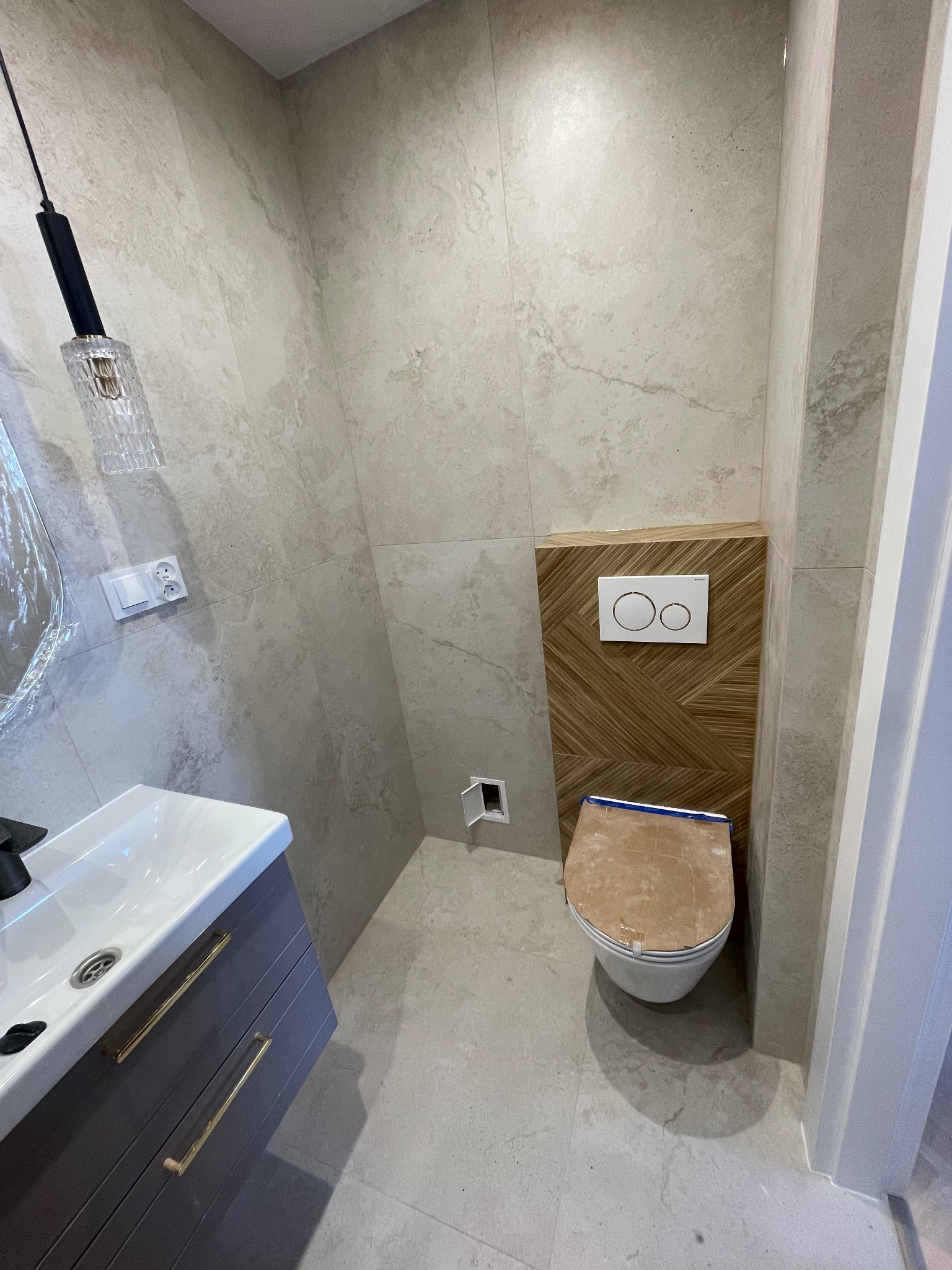 Nowoczesna łazienka w trakcie wykańczania: umywalka z szarą szafką, podwieszana toaleta z drewnianą obudową i płytki imitujące kamień na ścianach.