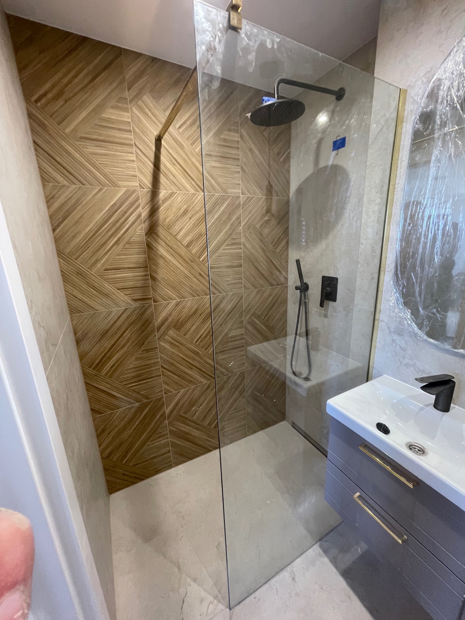 Nowoczesna łazienka z kabiną typu walk-in, drewnopodobnymi płytkami ułożonymi w geometryczny wzór i szarą szafką pod umywalką z złotymi uchwytami.