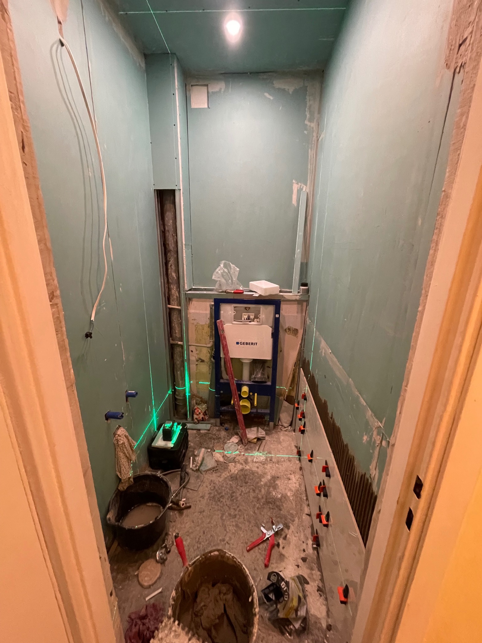 Wąska łazienka w trakcie remontu: widoczne płyty karton-gips, stelaż WC Geberit, poziomica laserowa, wiadro z zaprawą i fragment ułożonych płytek z krzyżykami dystansowymi.