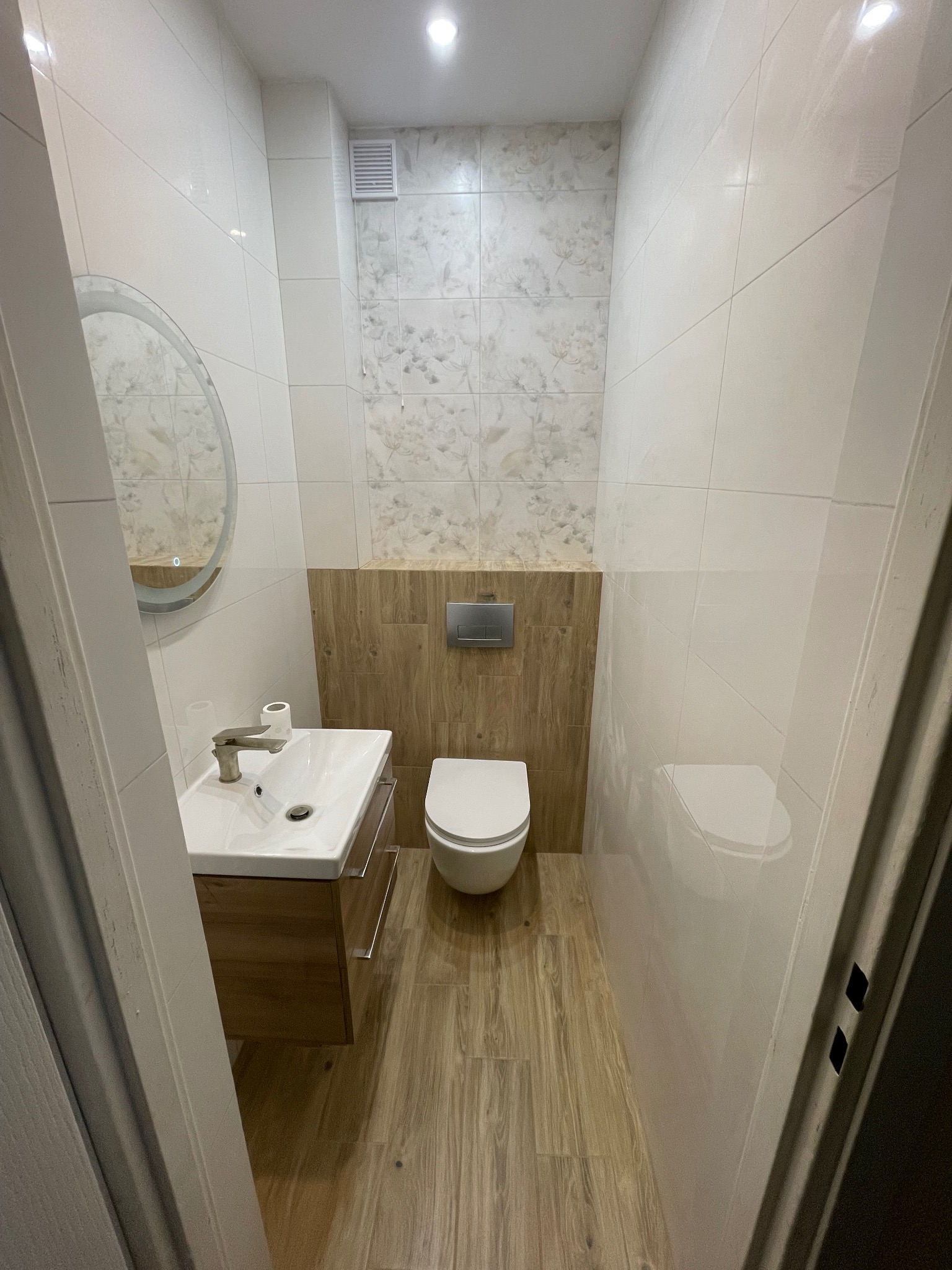 Nowoczesna łazienka po remoncie: białe płytki, drewnopodobna podłoga i zabudowa, okrągłe lustro z podświetleniem, toaleta i umywalka z szafką. Minimalistyczny design.