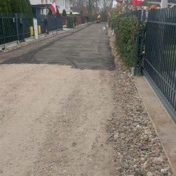 P.H.T.U - Fragment utwardzonej drogi osiedlowej z widocznymi polskimi flagami na posesjach. W tle maszyna budowlana i pies. Częściowo utwardzona nawierzchnia.