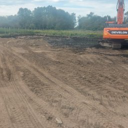 P.H.T.U - Pomarańczowa koparka Develon i zielona naczepa ciężarówki na placu budowy z wykopaną ziemią. Widoczne ślady opon i hałdy ziemi w tle.