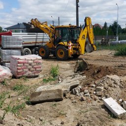 P.H.T.U - Żółta koparko-ładowarka JCB na placu budowy, obok zaparkowana ciężarówka z materiałami budowlanymi i stertą gruzu. Błękitne niebo z chmurami w tle.