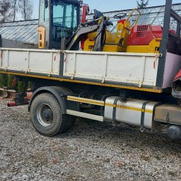 P.H.T.U - Ciężarówka z platformą załadowaną minikoparką i walcem drogowym na żwirowym podłożu, w tle szklarnia i niebo z chmurami.