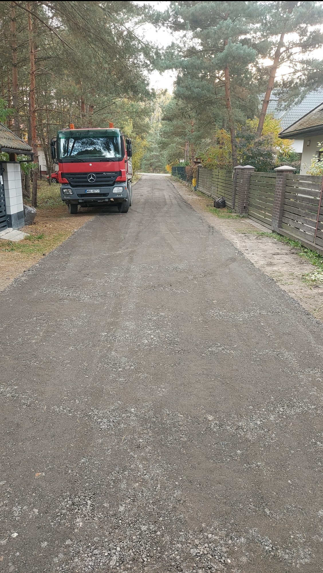 Ciężarówka Mercedes-Benz na świeżo utwardzonej, szarej drodze gruntowej, otoczonej drzewami i ogrodzeniami posesji. Widok z perspektywy drogi.
