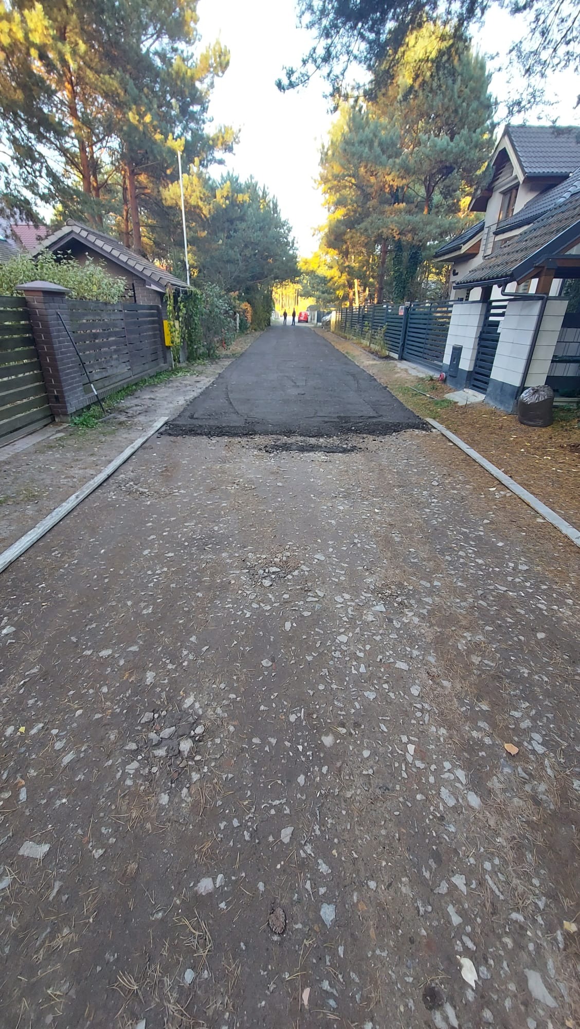Fragment utwardzonej drogi dojazdowej na osiedlu domów jednorodzinnych. Widoczne świeże asfaltowanie na tle istniejącej nawierzchni. Prace w toku.