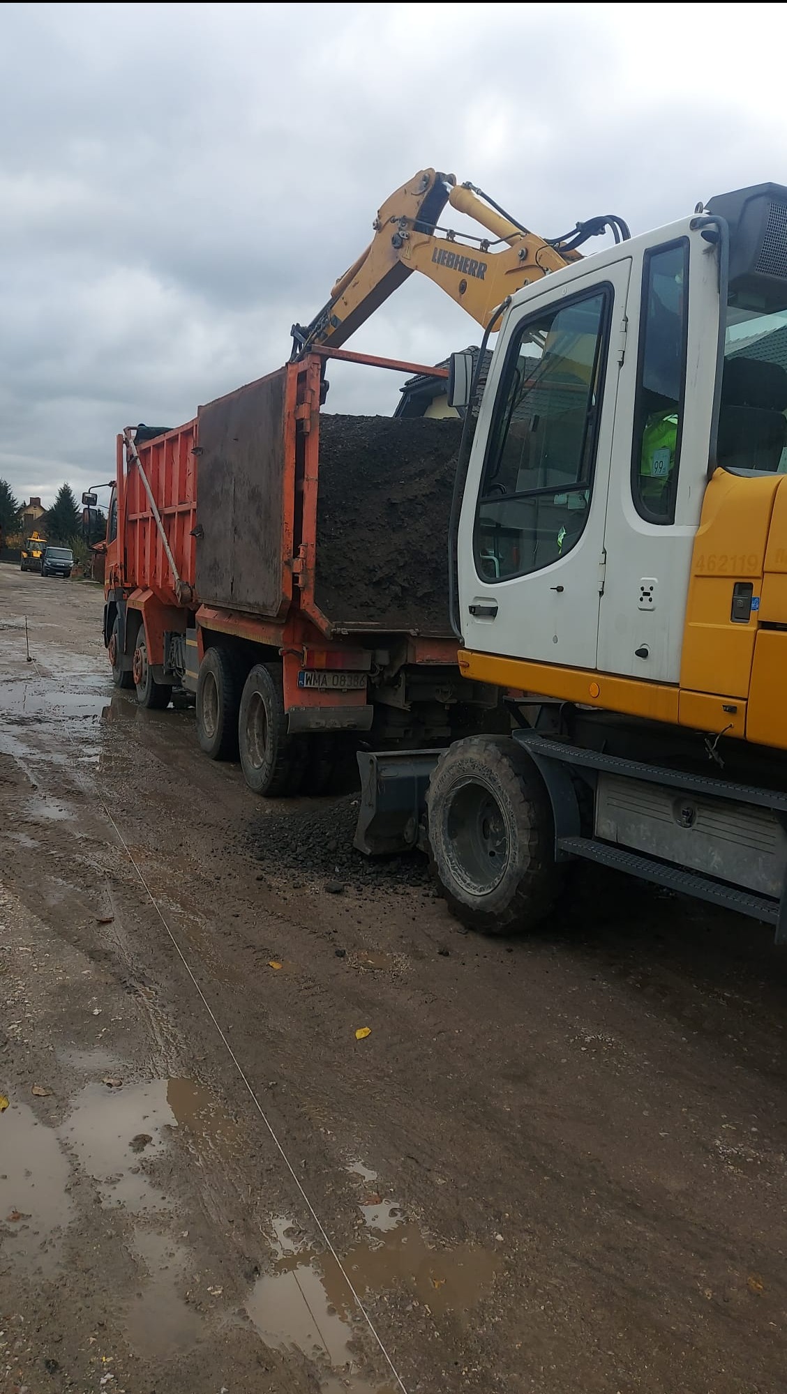 Żółta koparka Liebherr ładuje ziemię do pomarańczowej ciężarówki w Nieporęcie. Prace ziemne na błotnistym terenie, pochmurne niebo w tle.