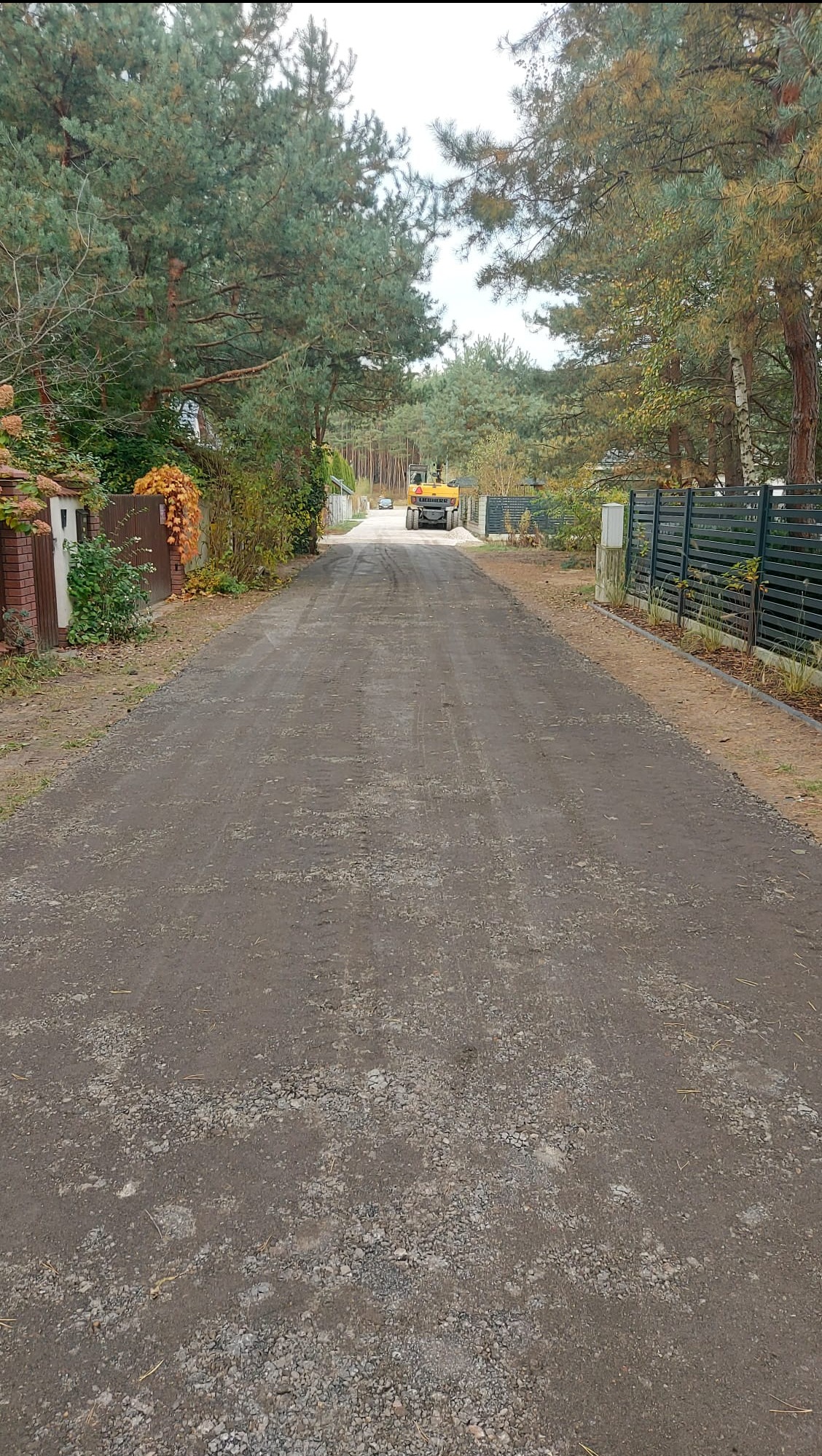 Utwardzona droga dojazdowa z walcem w tle, otoczona zielenią i ogrodzeniami. Widok perspektywiczny, podkreślający długość i gładkość nawierzchni.