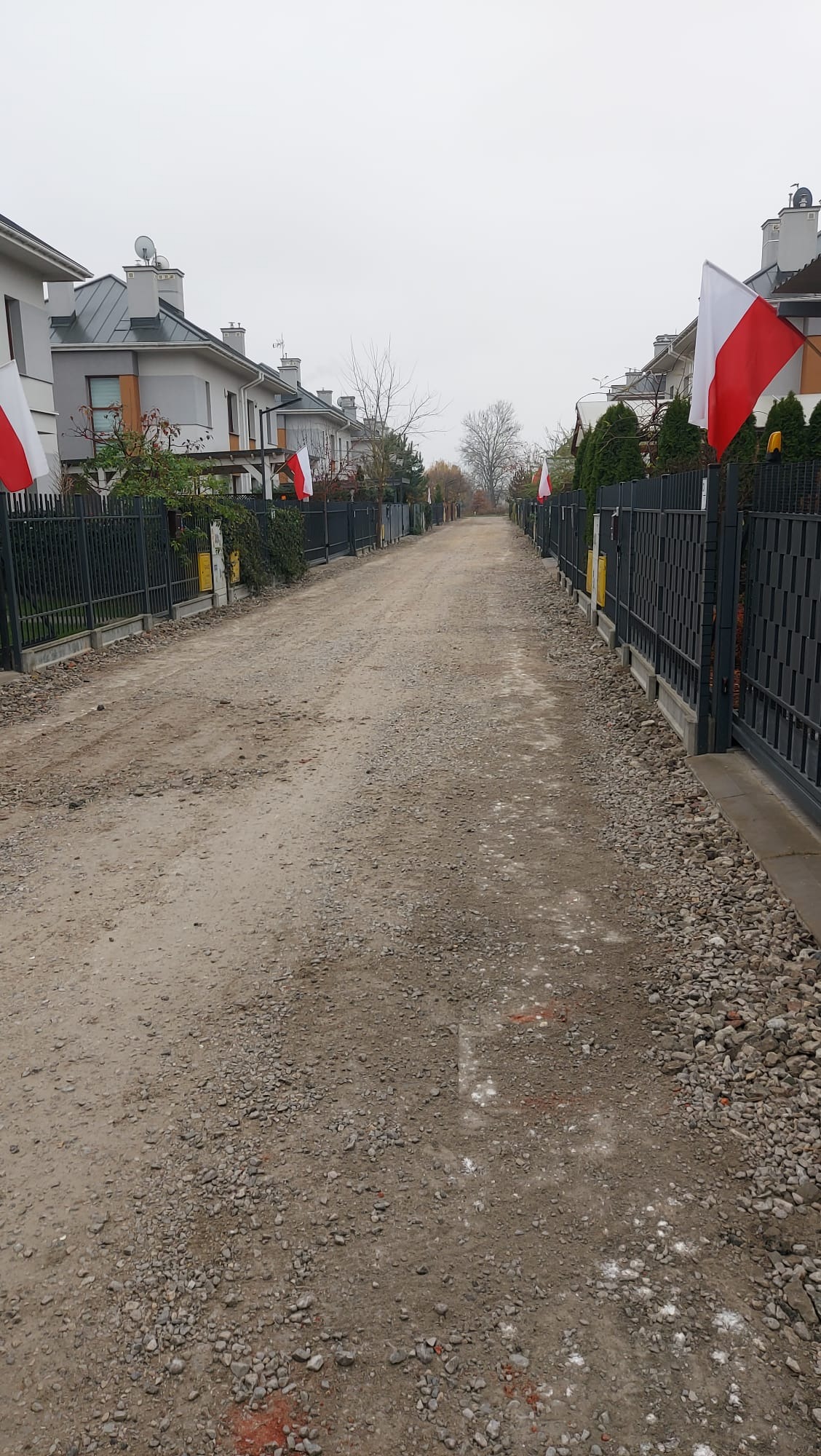 Ulica osiedlowa w trakcie utwardzania, widoczne domy z flagami Polski wzdłuż drogi. Szara, pochmurna pogoda.