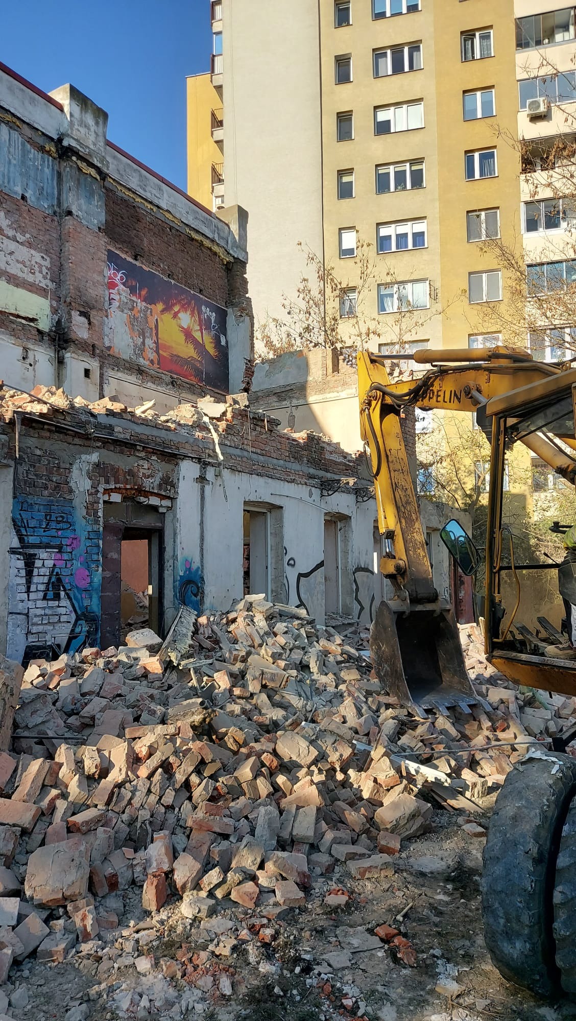 Żółta koparka na tle zrujnowanego budynku z cegły i graffiti. W tle bloki mieszkalne. Widoczne gruzy i pozostałości po rozbiórce.
