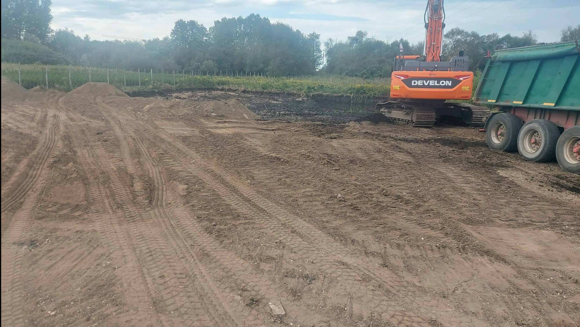 Pomarańczowa koparka Develon i zielona naczepa ciężarówki na placu budowy z wykopaną ziemią. Widoczne ślady opon i hałdy ziemi w tle.