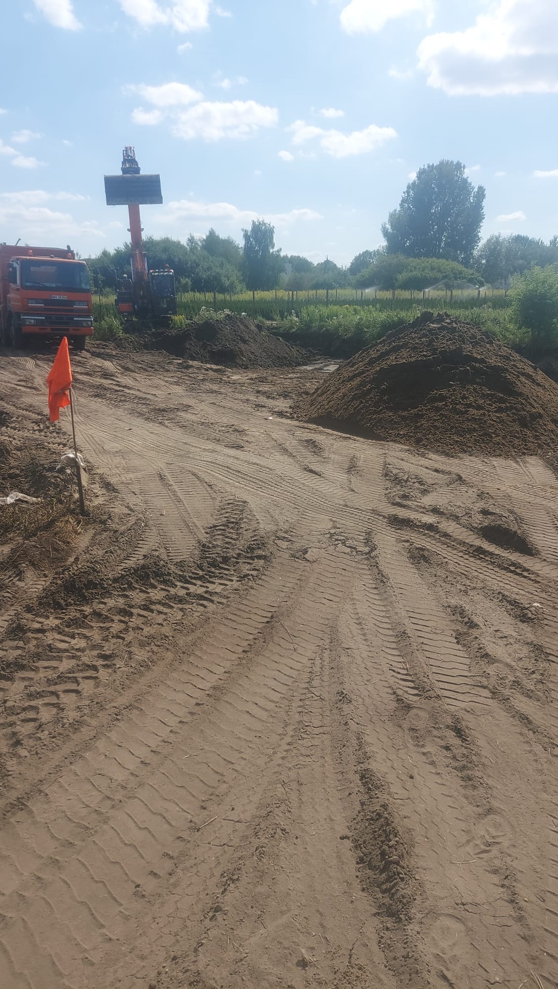 Praca ziemna w toku: koparka z łyżką, pomarańczowa ciężarówka i hałdy ziemi na placu budowy. Widoczne ślady opon i pomarańczowa chorągiewka.