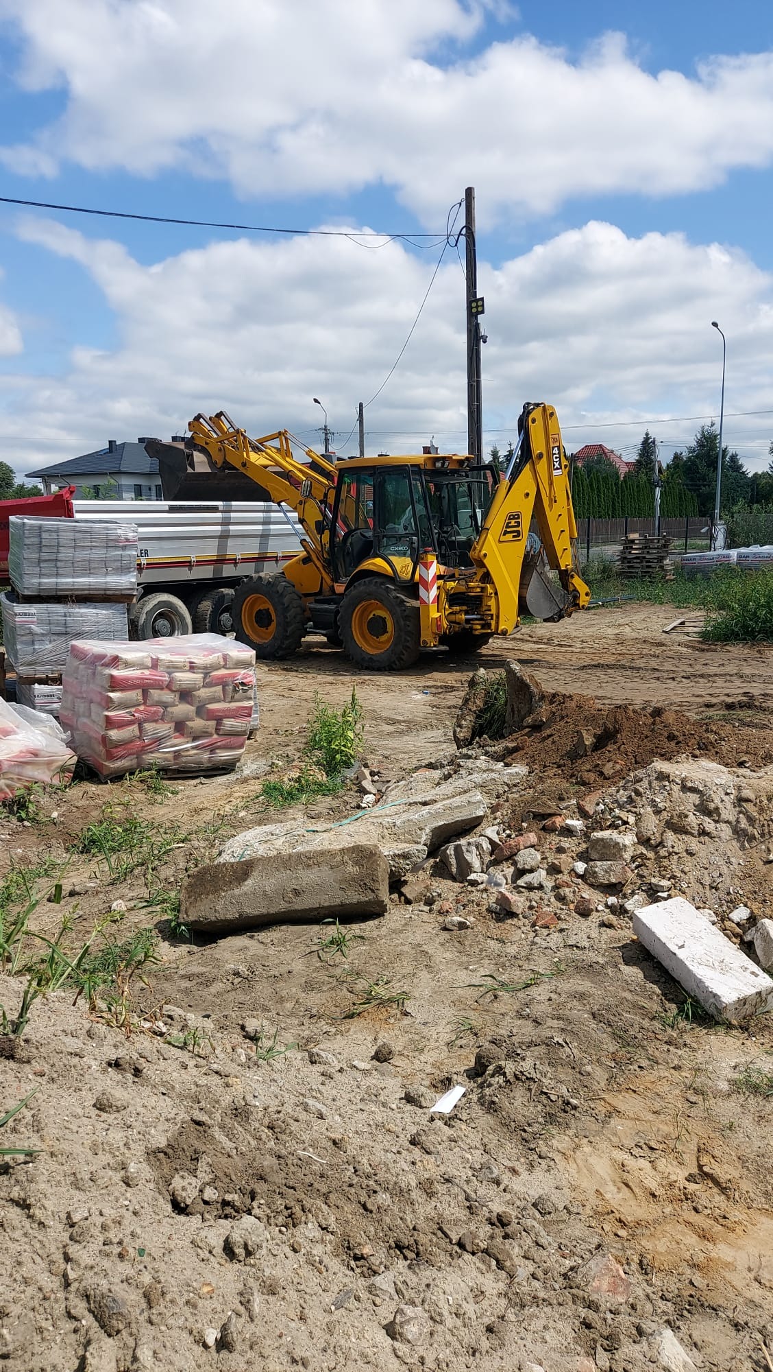 Żółta koparko-ładowarka JCB na placu budowy, obok zaparkowana ciężarówka z materiałami budowlanymi i stertą gruzu. Błękitne niebo z chmurami w tle.