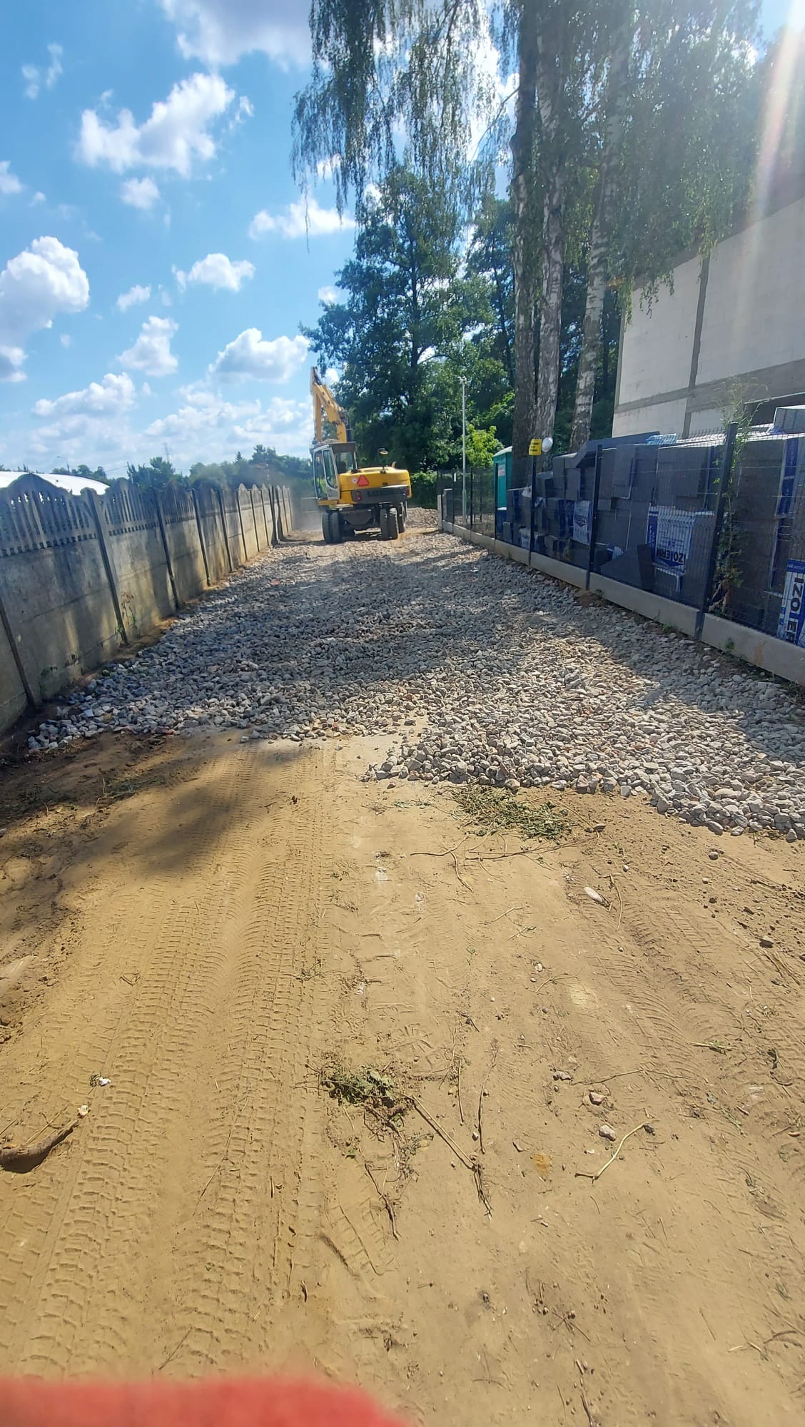 Żółta koparka na żwirowej drodze między betonowym ogrodzeniem a stertą materiałów budowlanych w słoneczny dzień. Widoczne ślady opon na piaszczystym podłożu.