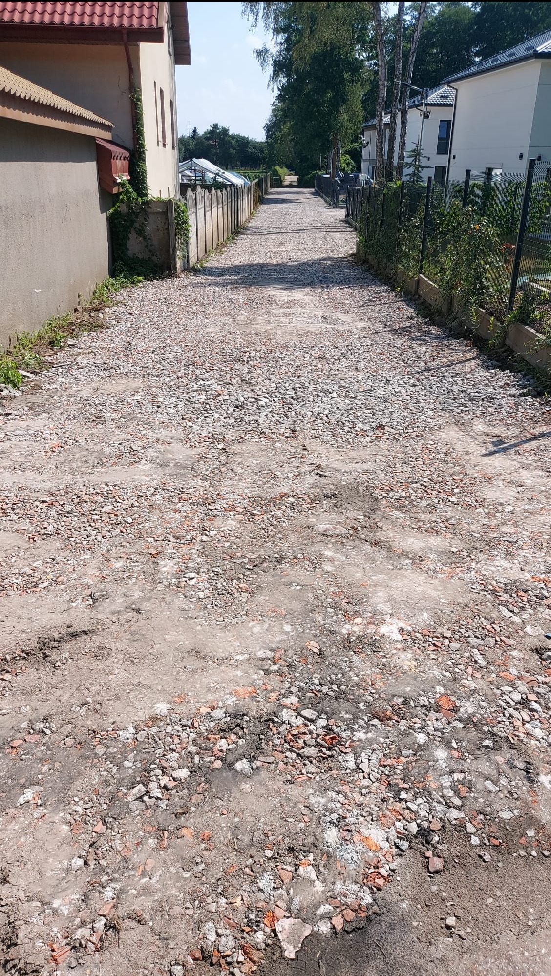 Ukończone utwardzanie drogi dojazdowej z gruzem i kamieniami, widok z perspektywy ulicy, domy po bokach, słoneczny dzień.