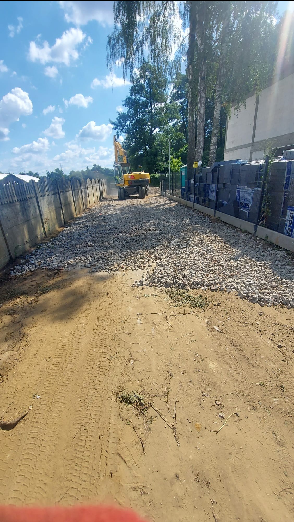Żółta koparka na żwirowej drodze między betonowym ogrodzeniem a paletami z materiałami budowlanymi, błękitne niebo z chmurami w tle.