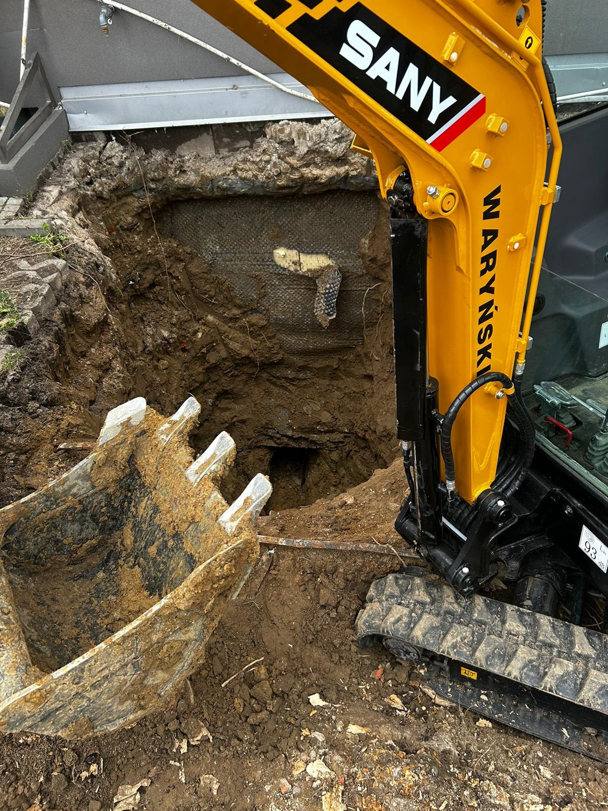 Żółta koparka SANY z łyżką w ziemi, widoczny wykop obok budynku. Prace ziemne, wykop pod instalacje.