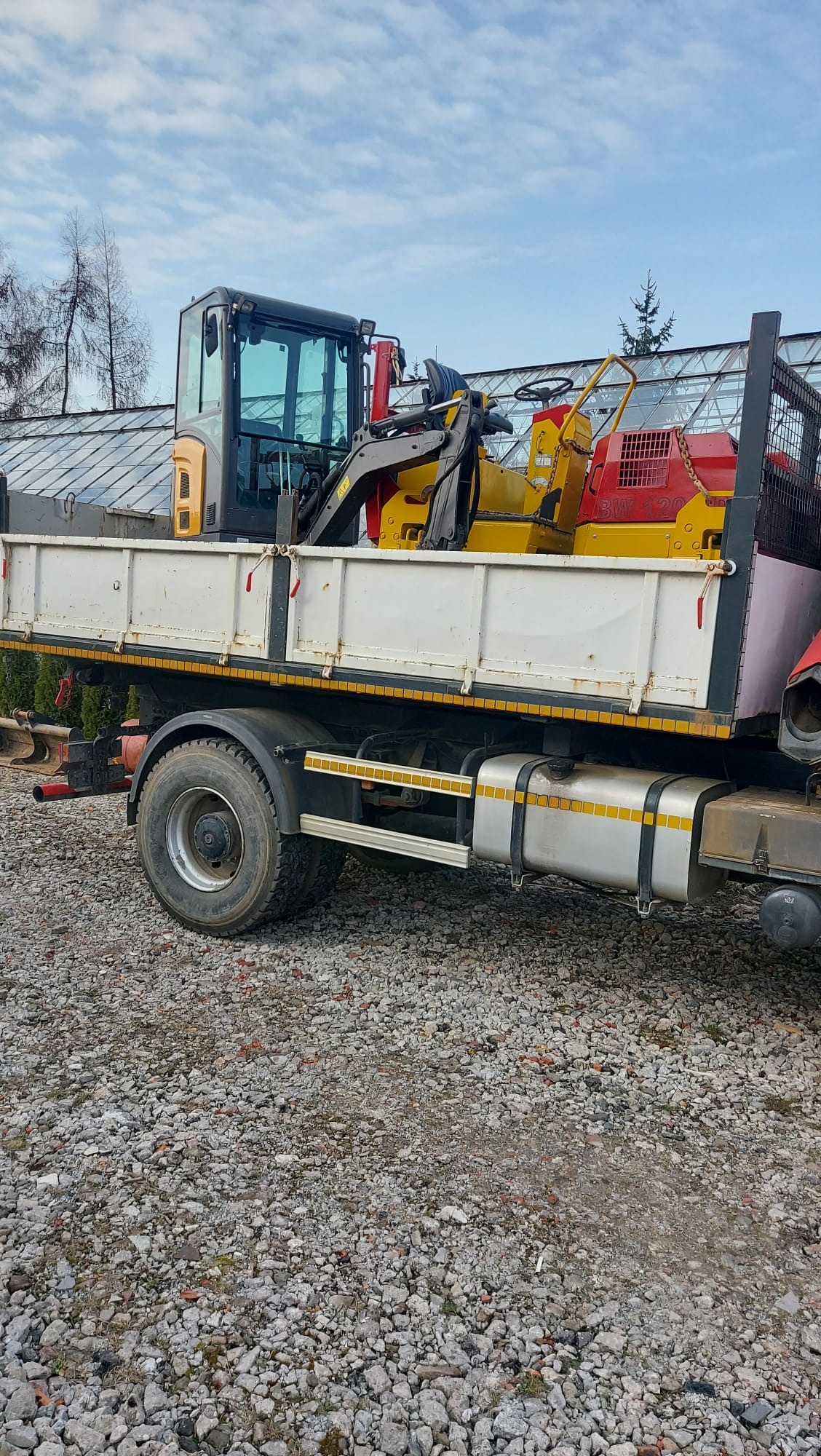 Ciężarówka z platformą załadowaną minikoparką i walcem drogowym na żwirowym podłożu, w tle szklarnia i niebo z chmurami.