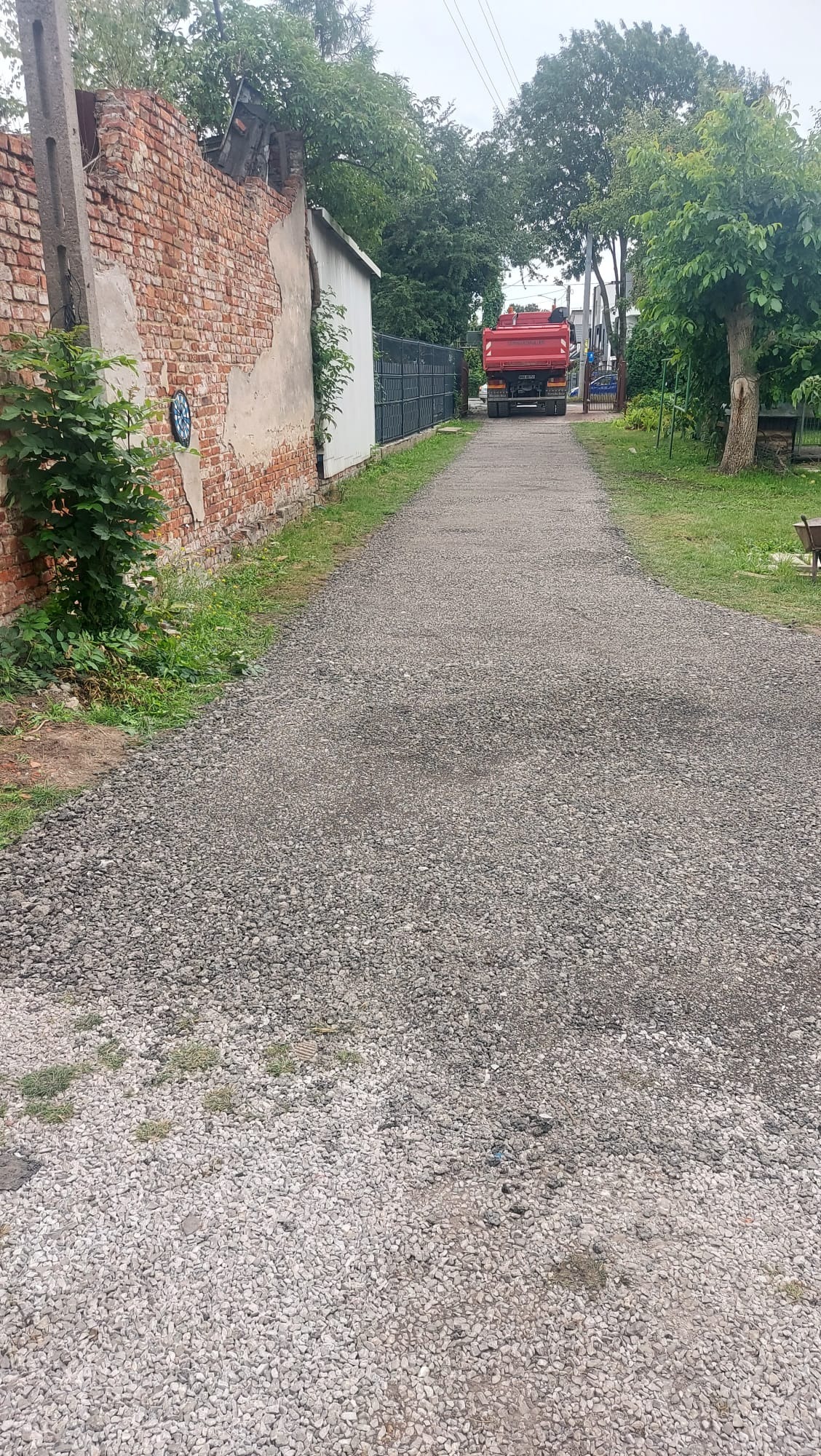 Utwardzona droga z szarego kruszywa prowadząca w kierunku czerwonej ciężarówki. Po bokach widoczne fragmenty muru i zieleni.