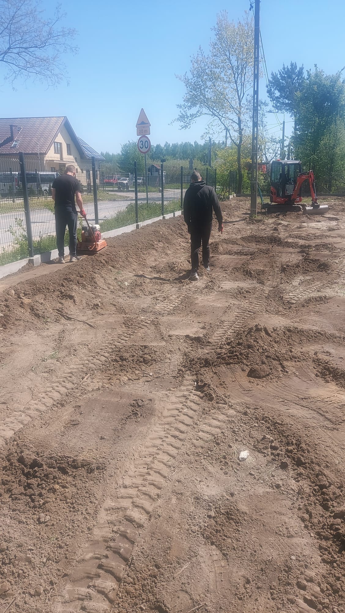 Prace ziemne: ubijanie gruntu zagęszczarką wzdłuż ogrodzenia. W tle minikoparka i pracownik. Ślady opon na ziemi. Słoneczny dzień.