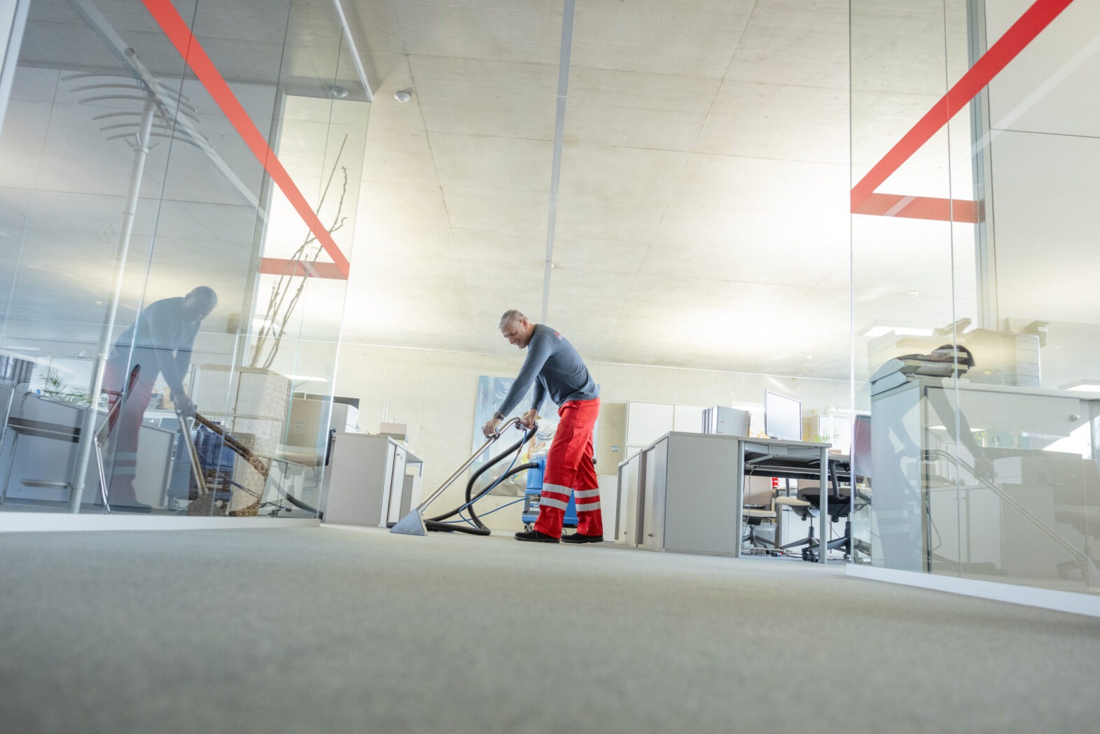 Pracownik w czerwonych spodniach czyści wykładzinę w biurze za pomocą profesjonalnego odkurzacza piorącego. Widoczne szklane ściany działowe i biurka.