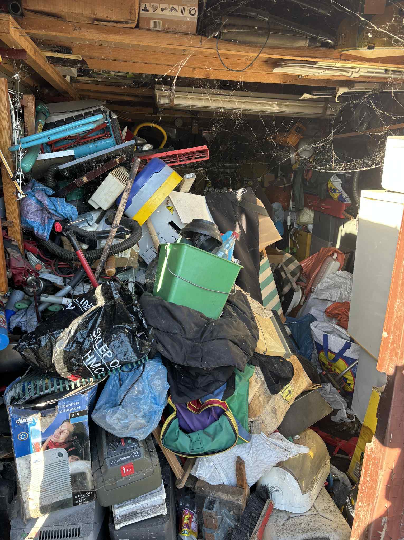 Zapełnione rupieciami pomieszczenie z pajęczynami pod sufitem. Widoczne worki, pudełka, sprzęty i inne przedmioty w nieładzie, tworzące wrażenie zagraconej przestrzeni.