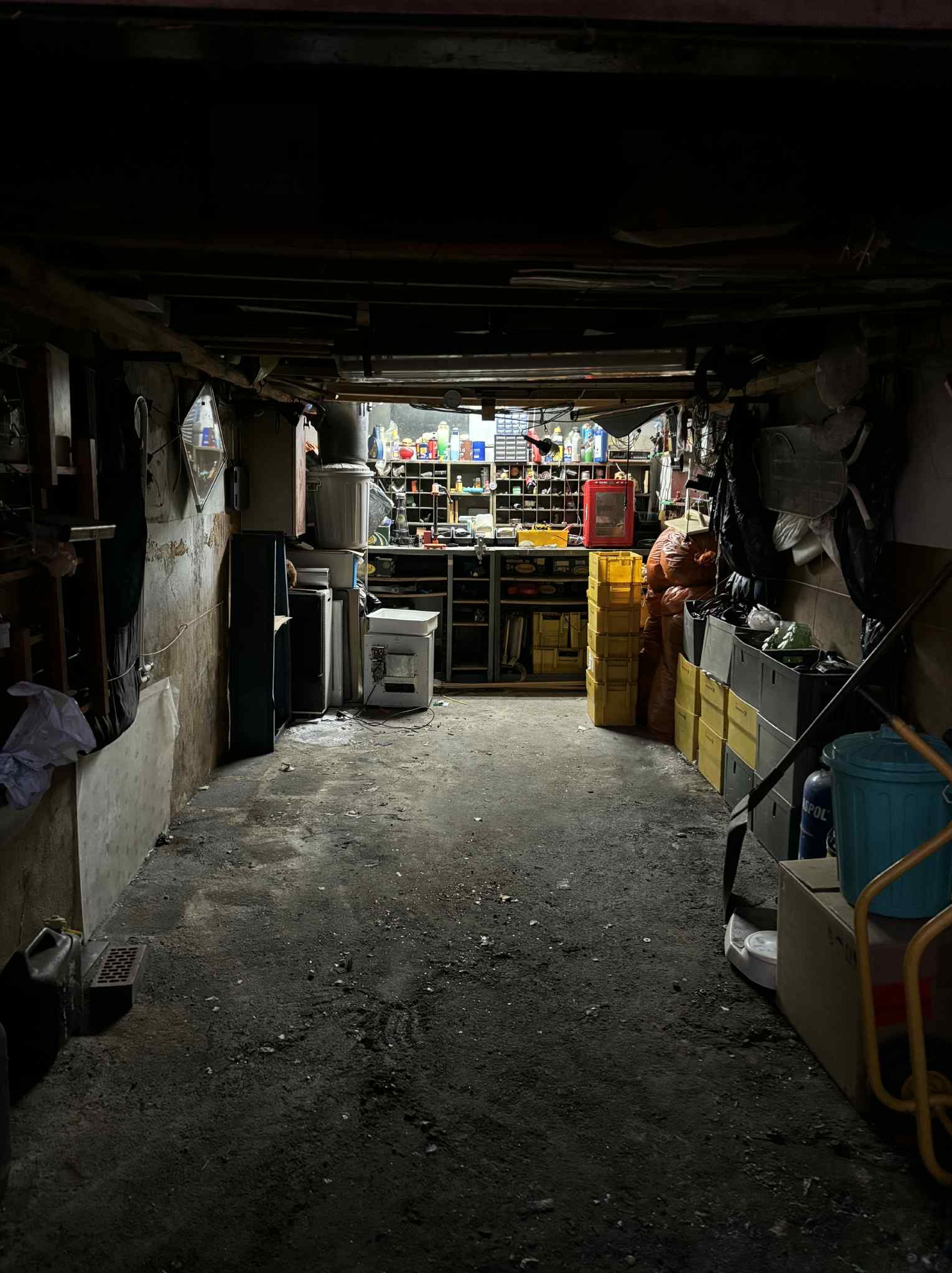 Zagracony garaż lub piwnica z narzędziami, pojemnikami i sprzętami. Widoczne regały, skrzynki, butle i inne przedmioty. Nierówna podłoga, słabe oświetlenie.