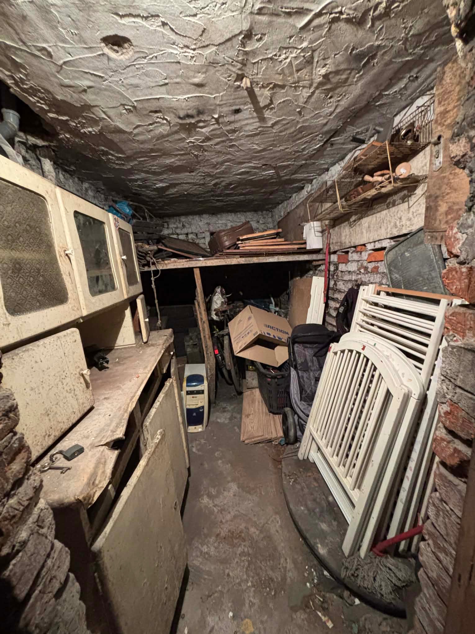 Zagracone pomieszczenie z meblami, rowerem i kartonami. Stare szafki, ceglana ściana i sufit z nierówną fakturą. Widoczny bałagan i przedmioty przeznaczone do usunięcia.