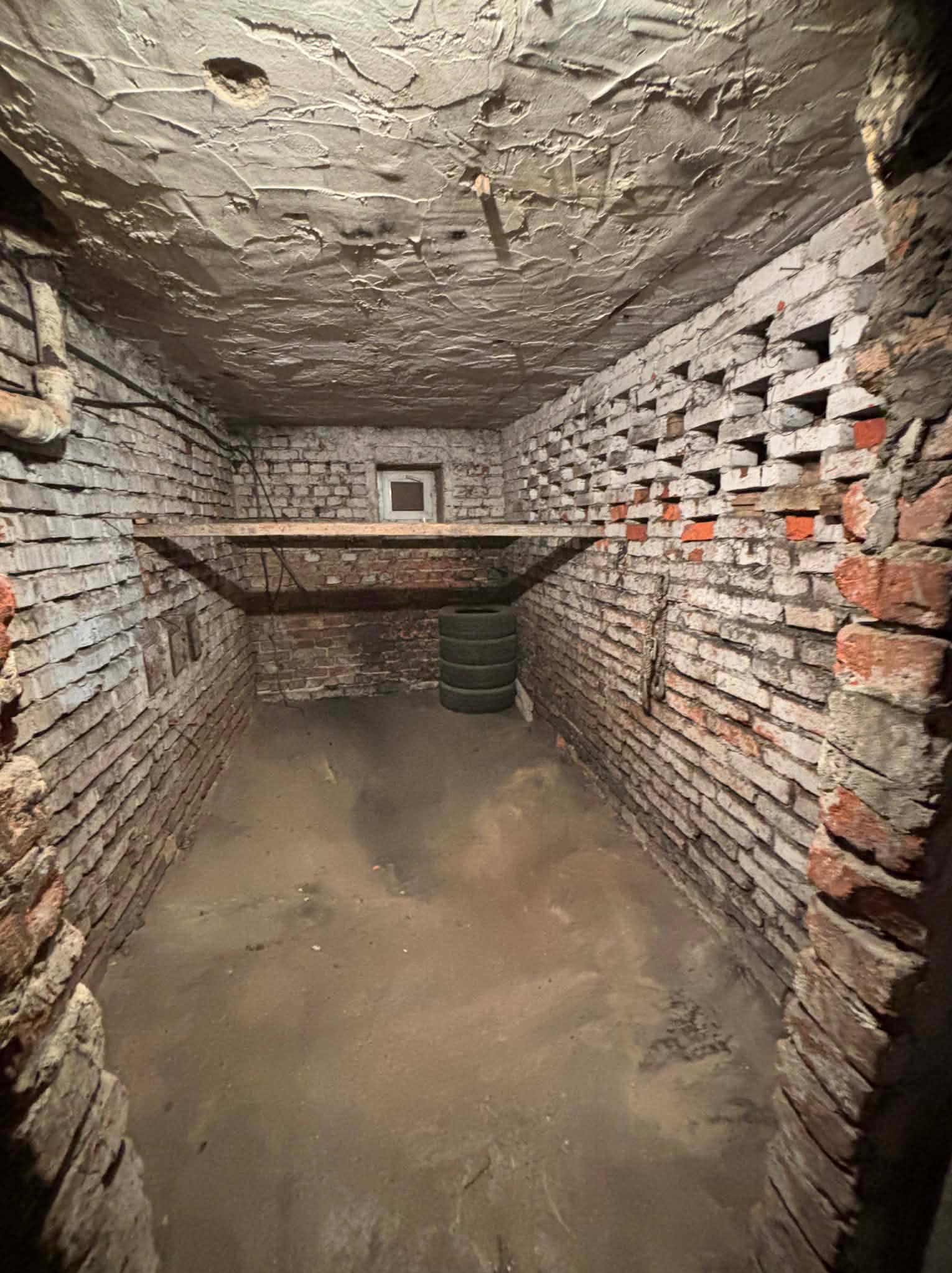 Ciemne, ceglane wnętrze piwnicy w Radomsku z drewnianą półką i oknem. Widoczne opony w rogu. Ściany z nierówną fakturą, surowy klimat.