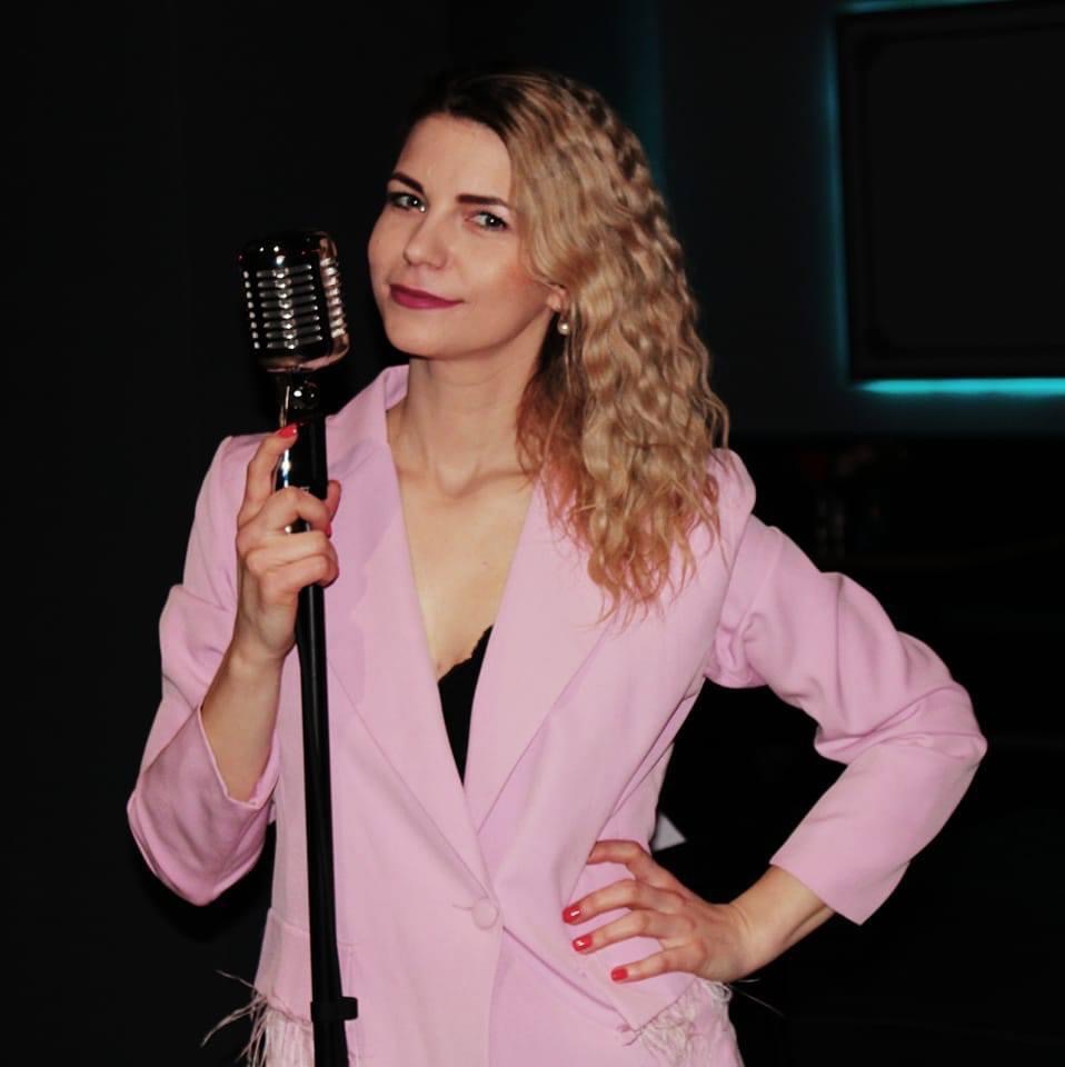 Uśmiechnięta blondynka w różowym żakiecie pozuje z mikrofonem retro na tle ciemnej sceny z subtelnym, niebieskim oświetleniem.