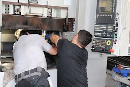 Dwóch serwisantów naprawia maszynę CNC, jeden z kluczem nasadowym, drugi przy panelu sterowania. Widoczne narzędzia i elementy maszyny. Jasne oświetlenie.