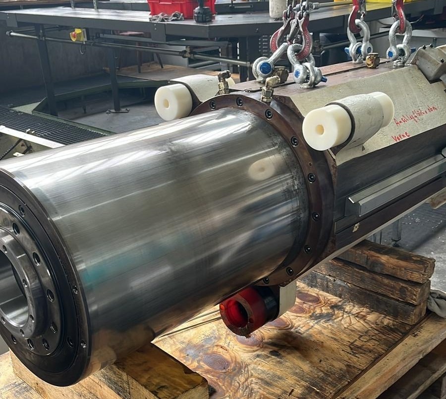 Polerowany, metalowy cylinder maszyny zawieszony na hakach, widoczne ślady użytkowania i napisy. Cylinder spoczywa na drewnianej palecie, w tle warsztat.