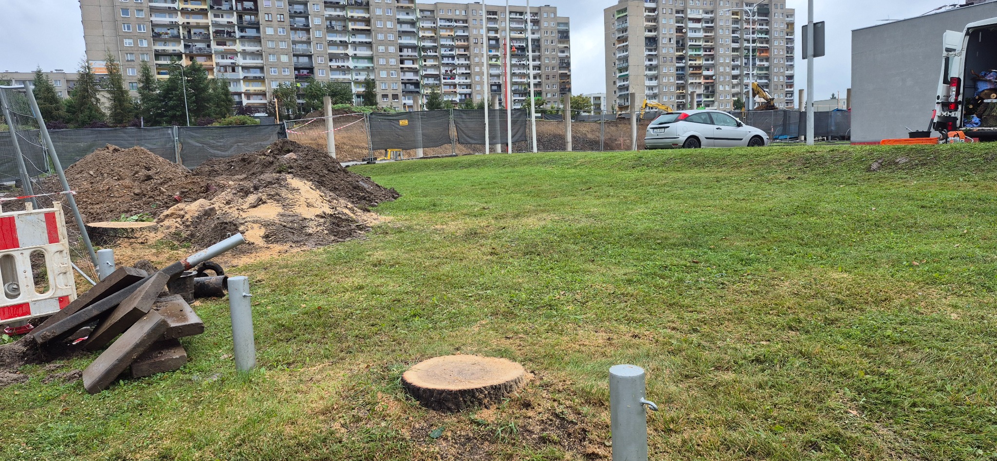 Świeżo ścięty pień drzewa na trawniku, w tle wykop, ogrodzenie i bloki mieszkalne. Widoczny samochód i narzędzia. Prace ziemne w przestrzeni miejskiej.