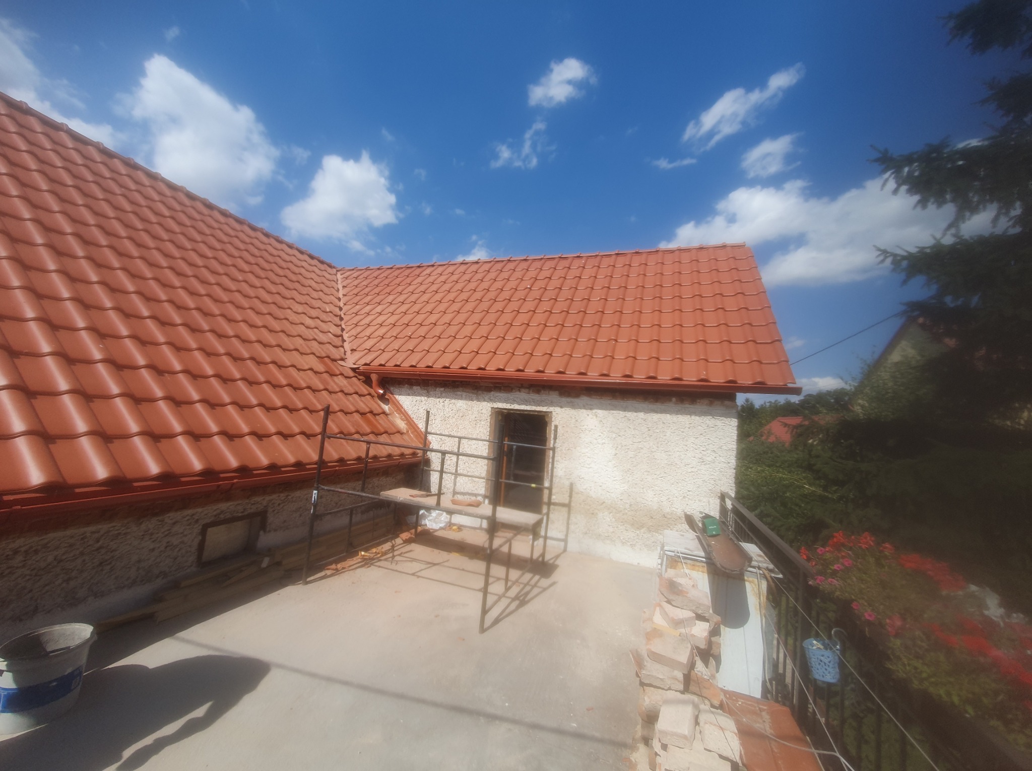 Czerwony dach z dachówki ceramicznej na budynku z rusztowaniem. Widok z góry na fragment dachu i elewacji w słoneczny dzień.