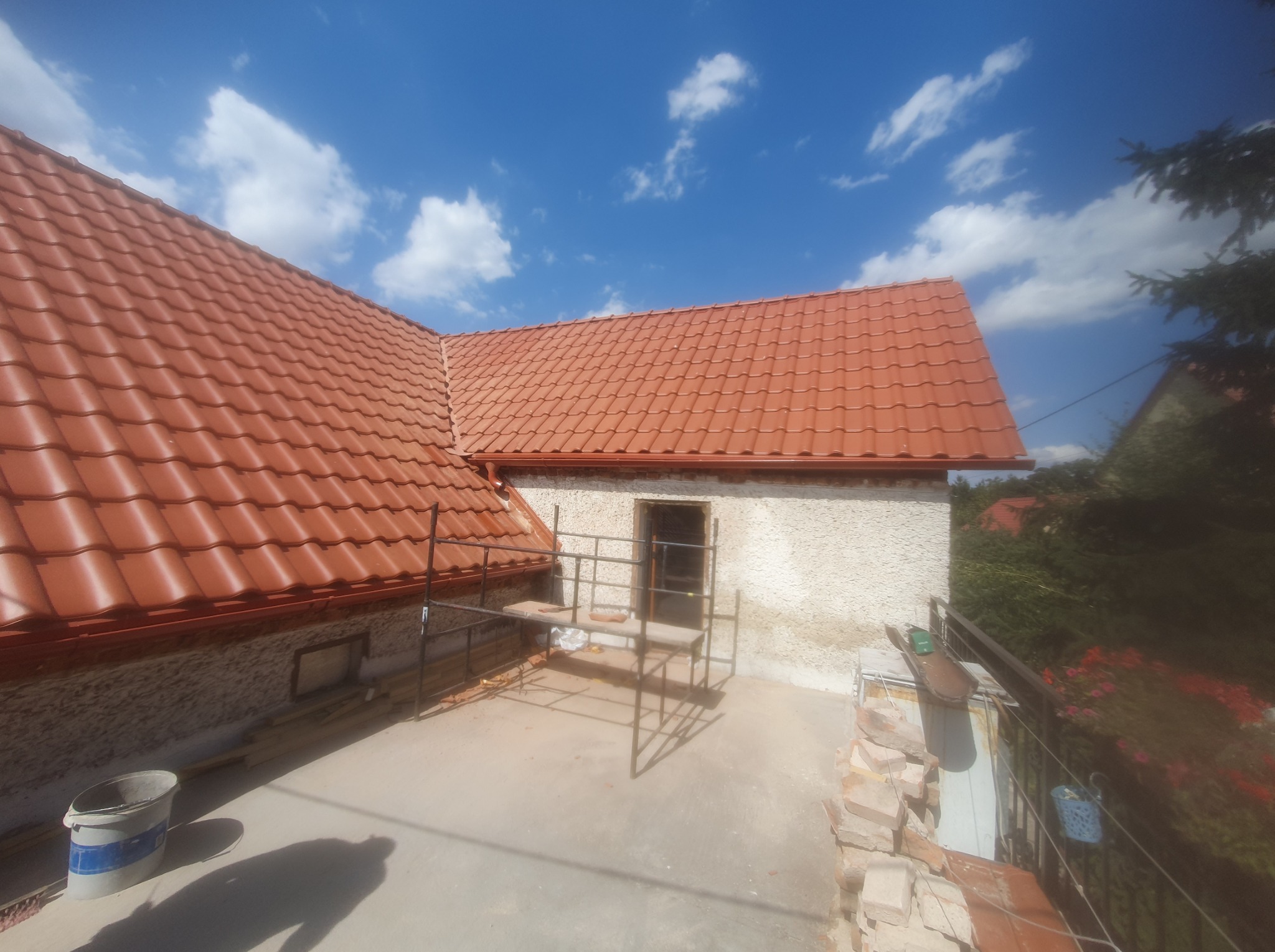 Czerwony dach z blachodachówki, widok z góry na rusztowanie i fragment elewacji. Błękitne niebo z chmurami w tle. Prace dekarskie w toku.