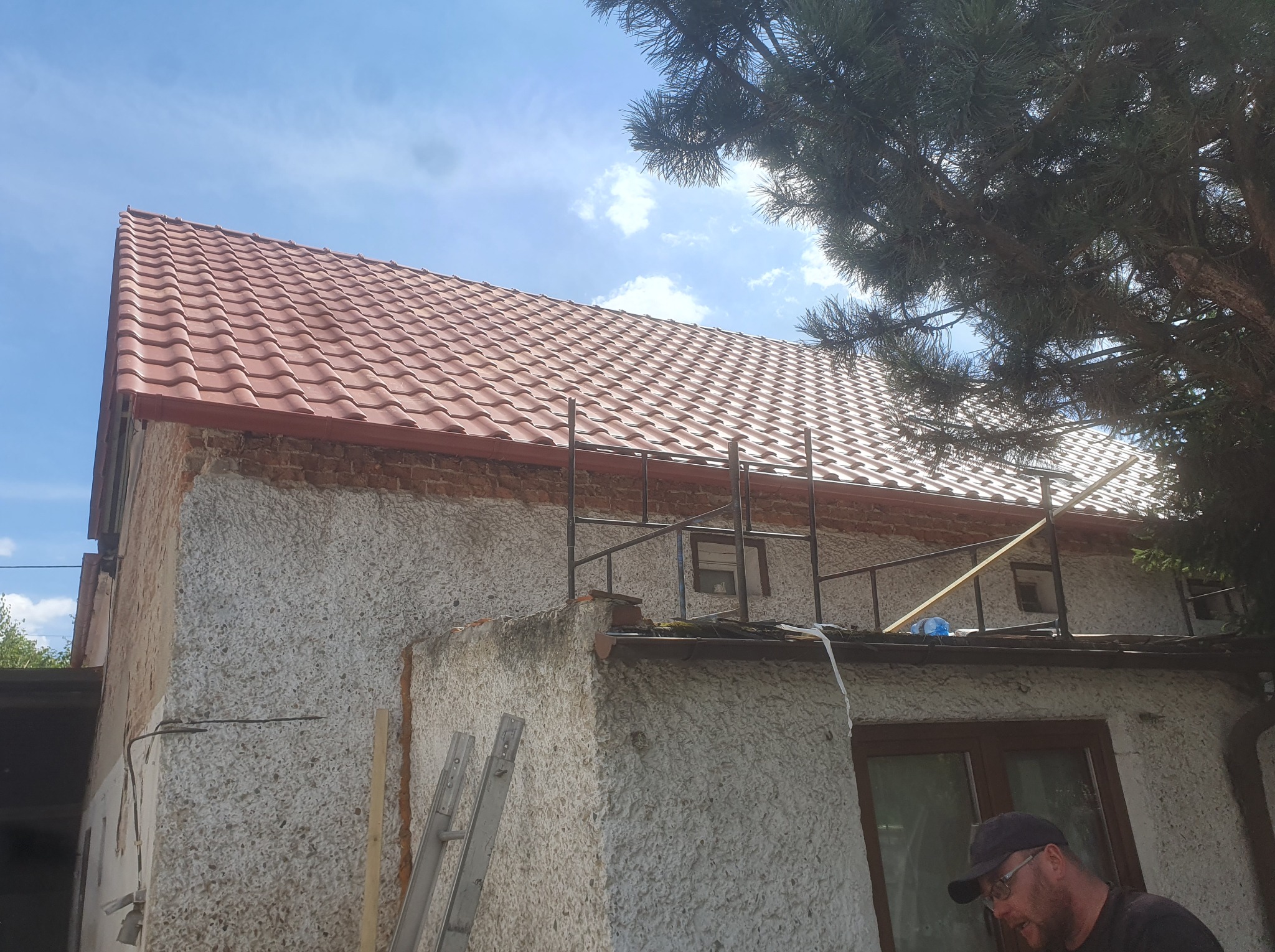 Ceglasty dach z nową dachówką i rynnami na budynku w Nysie, widok z dołu. Fragment rusztowania i drabina oparta o ścianę. Prace dekarskie w toku, widoczne detale wykończenia.