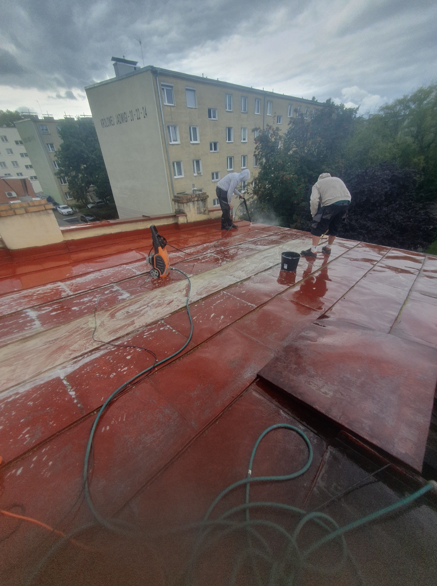 Mycie dachu z blachy na budynku mieszkalnym. Dwóch pracowników używa myjek ciśnieniowych. Widoczne ślady czyszczenia i budynek w tle.