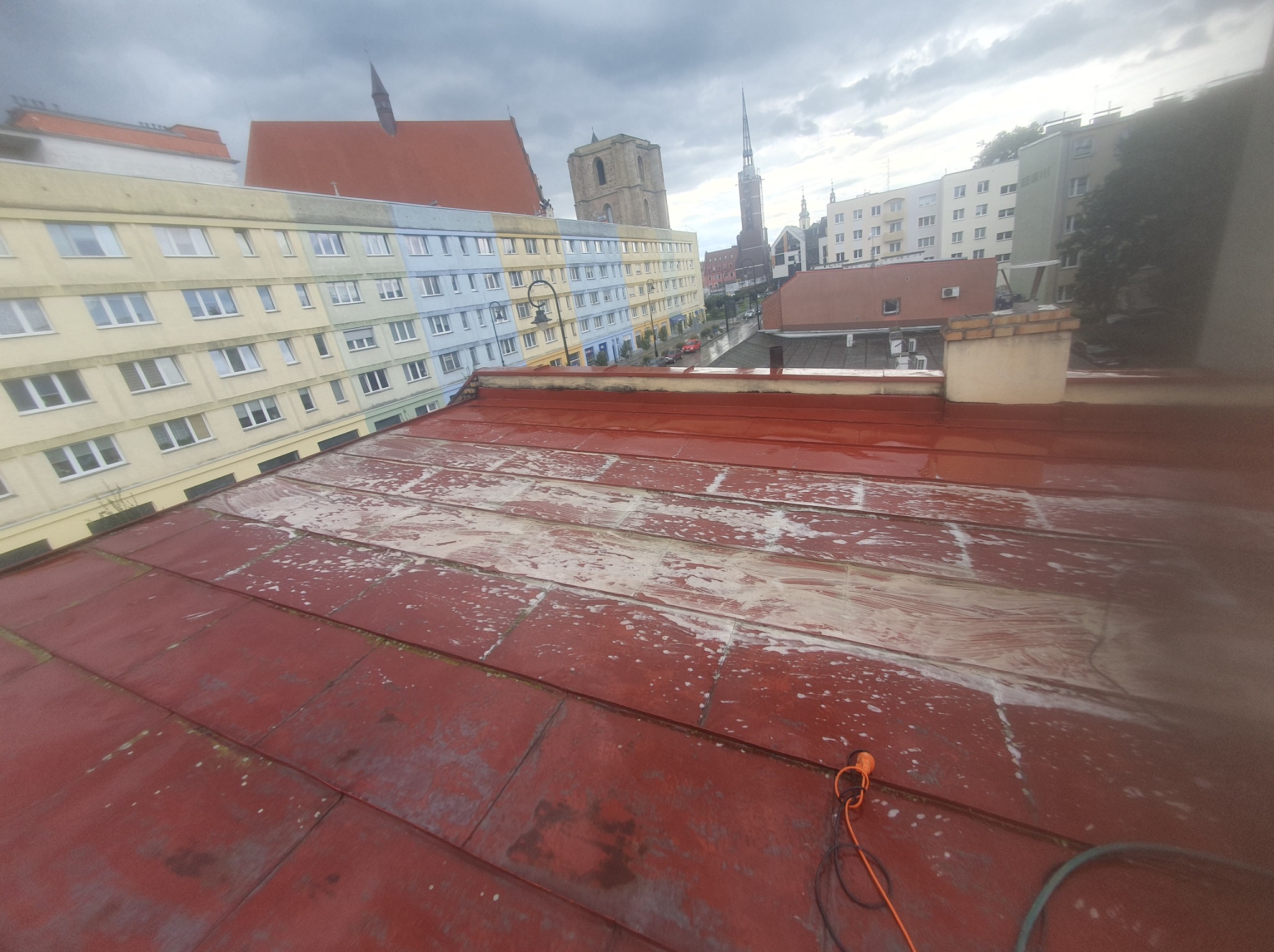 Czerwony dach z blachy, częściowo oczyszczony, z widocznym narzędziem i przewodem. W tle budynki miejskie i pochmurne niebo. Prace dekarskie w toku.