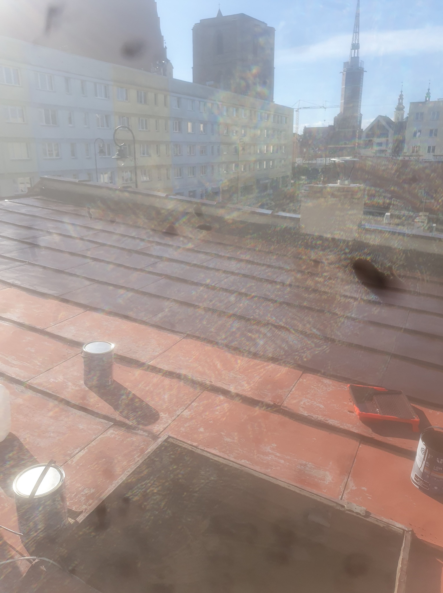 Malowanie dachu z blachy trapezowej na ceglasty kolor. Widoczne puszki z farbą i wałek. W tle zabudowa miejska i błękitne niebo. Praca w toku.