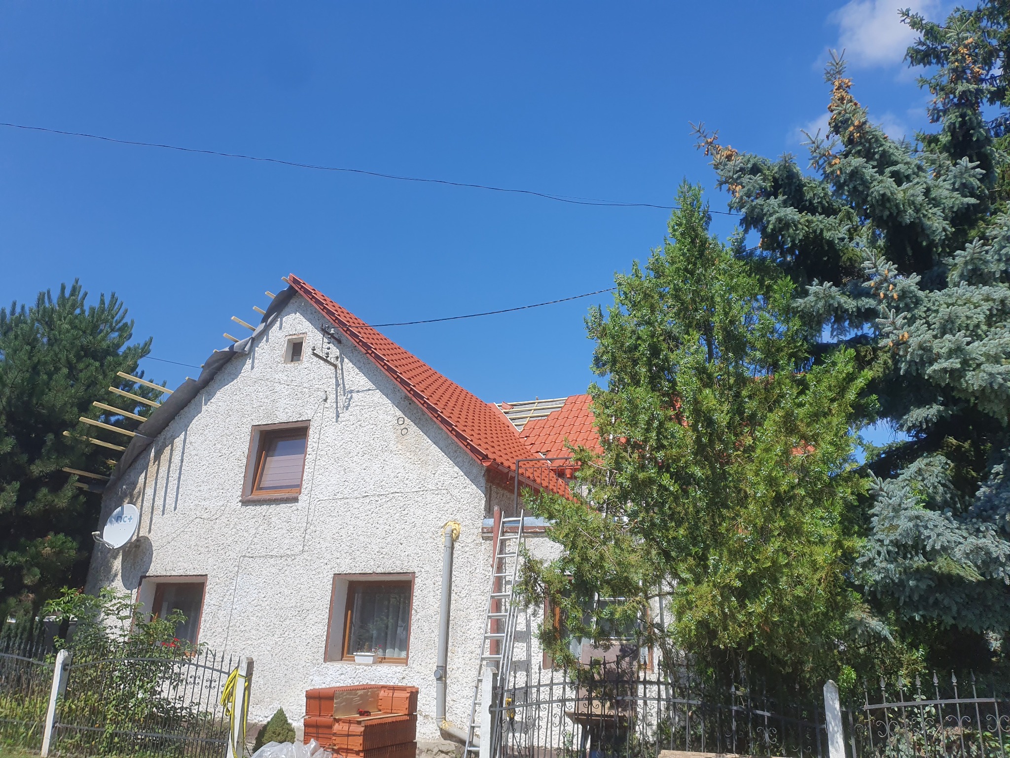 Dom z częściowo zdjętym dachem, widoczne krokwie i nowa dachówka. Przystawiona drabina, obok drzewa. Fragment elewacji z fakturą baranka.