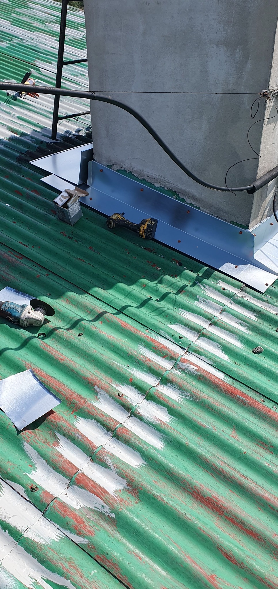 Zielony dach z blachy trapezowej w trakcie obróbek blacharskich komina. Widoczna wkrętarka, puszka z wkrętami i szlifierka kątowa.